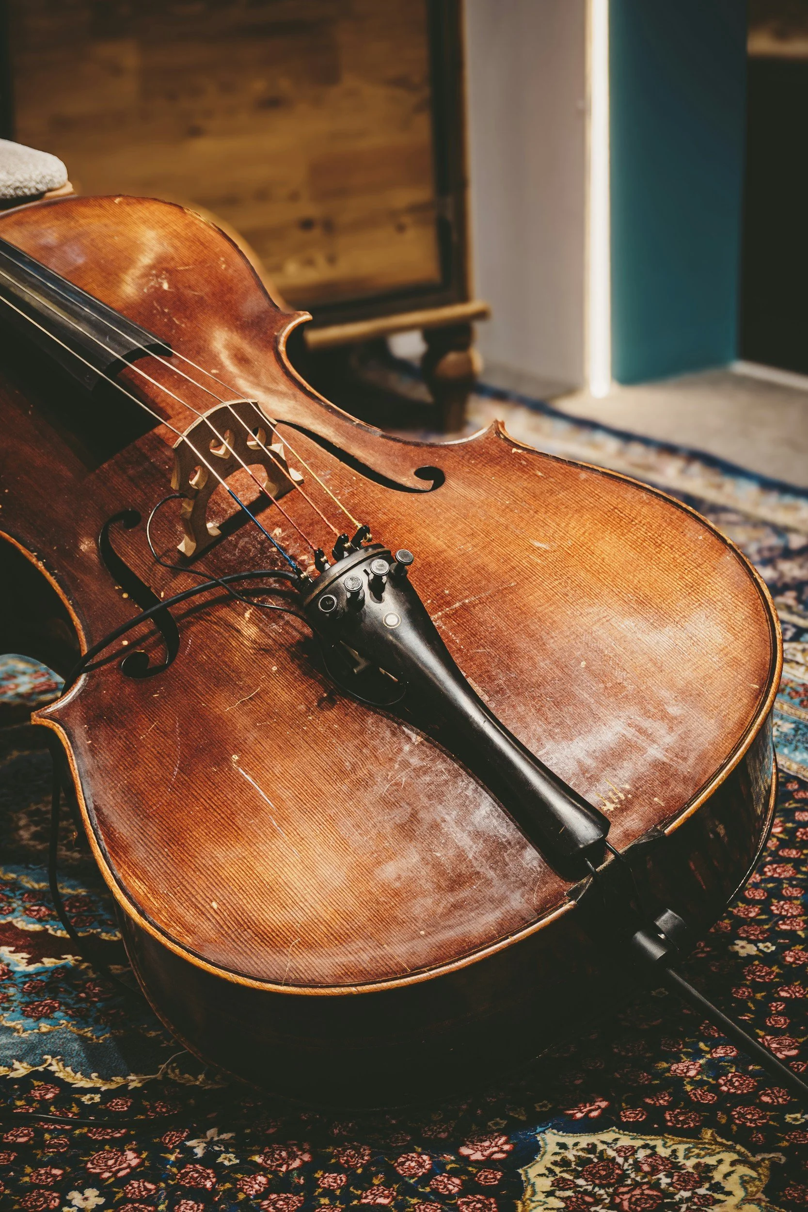 Close-up of an old wooden acoustic violin with visible wear, resting on a patterned rug, with a blurred background of furniture and a doorway.