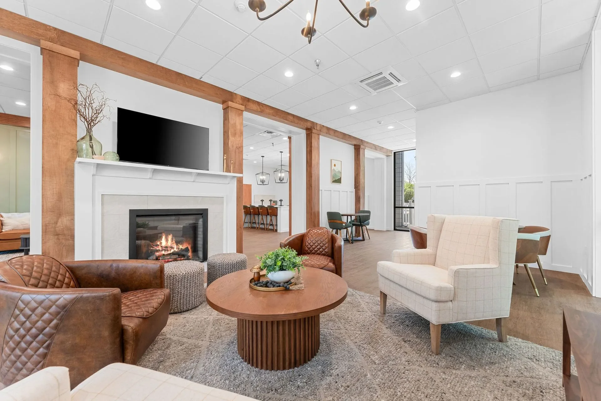 Cozy living room with a fireplace, a flat-screen TV above the mantel, a round wooden coffee table with a plant, armchairs, and a plush carpet, with a view of a dining area and large windows bringing in natural light.