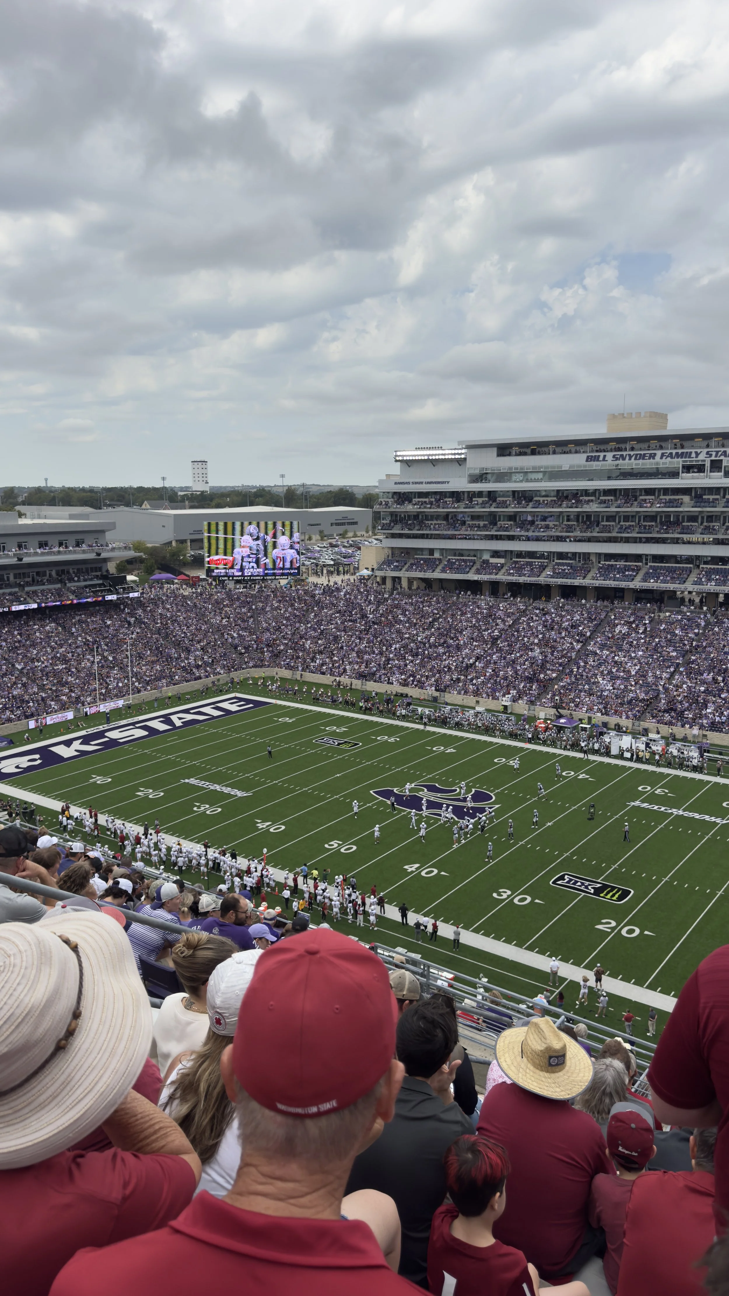 A football stadium filled with spectators watching a game. The players are on the field, and a large screen shows a close-up of players in uniform. The sky is cloudy.