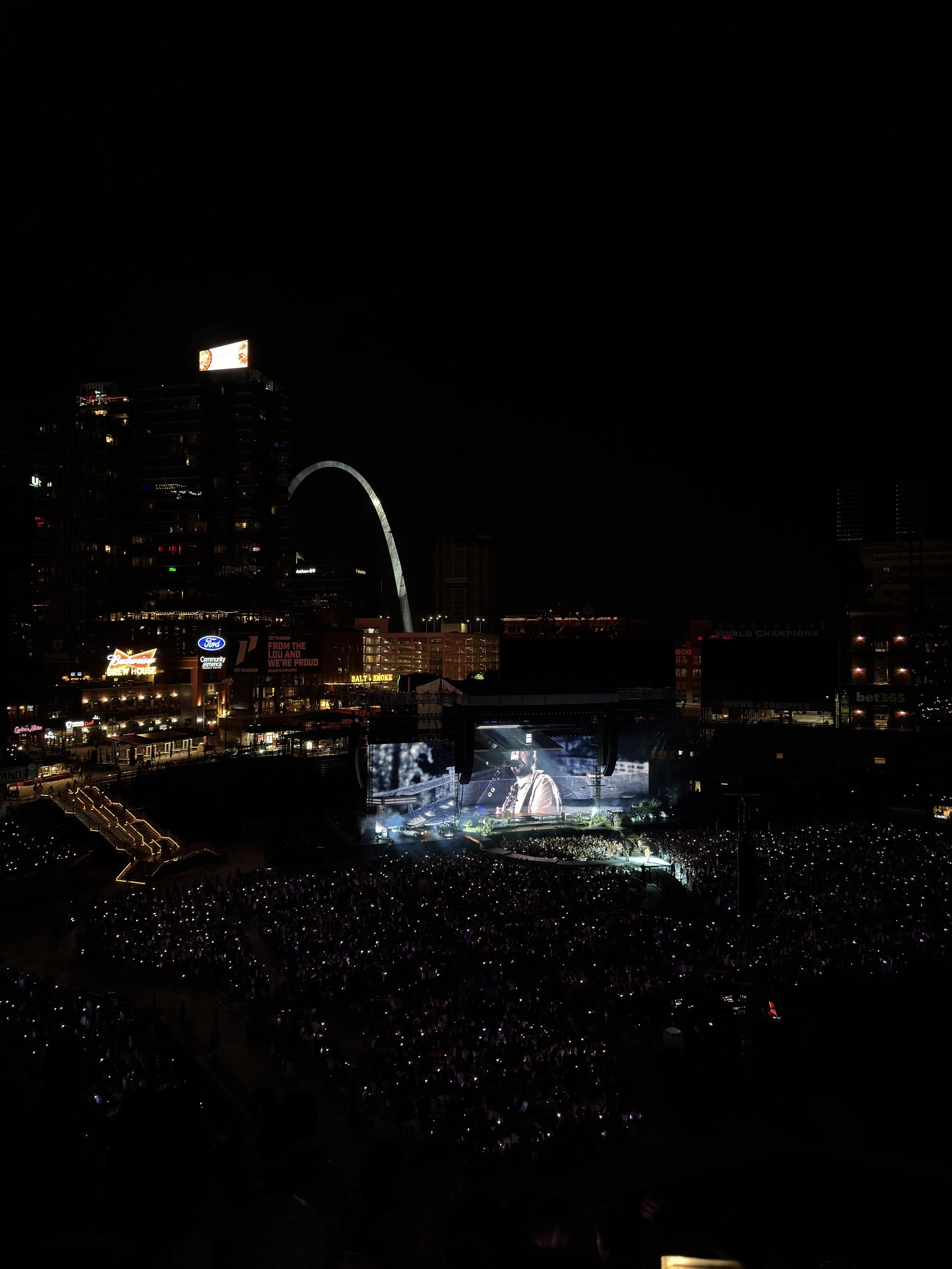 Nighttime concert in a city skyline with illuminated buildings and a large crowd watching a stage performance.