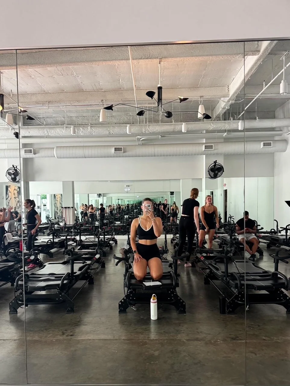 Mirror selfie of a woman in workout attire at a fitness class studio, with other people and exercise equipment in the background.