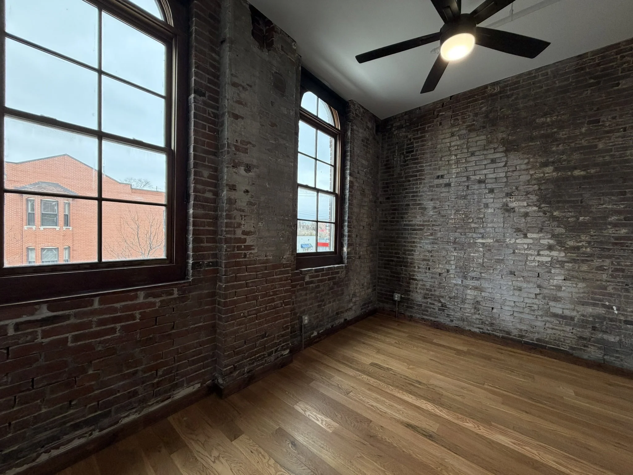 An empty room with exposed brick walls, large windows, a ceiling fan with light, and wooden floors.
