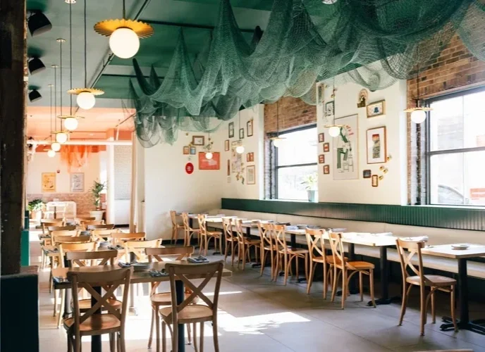 Empty restaurant with wooden chairs and tables, green ceiling drapes, and framed art on white walls.