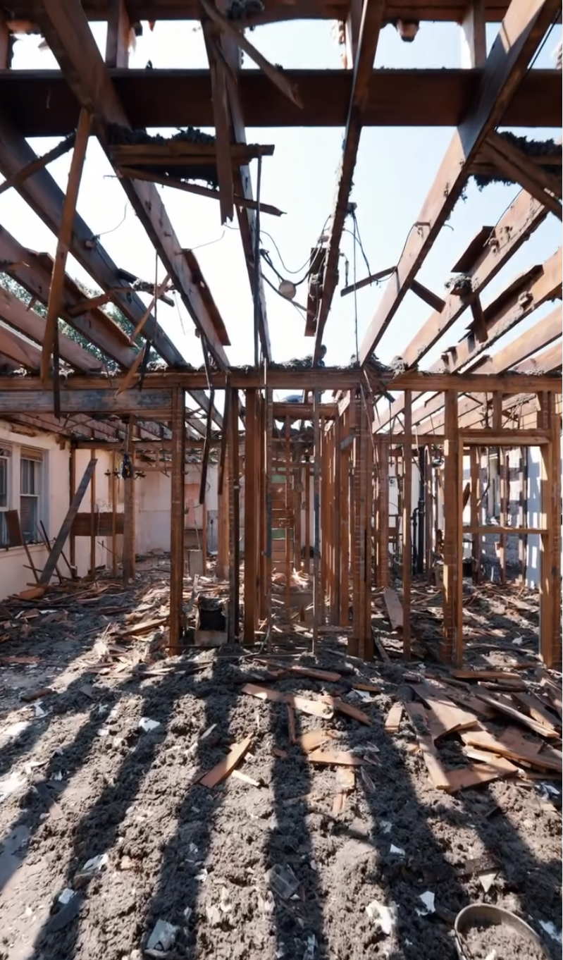 The interior of a building under demolition or construction, with wooden framing and exposed roof structure, debris on the ground, and debris hanging from the ceiling.
