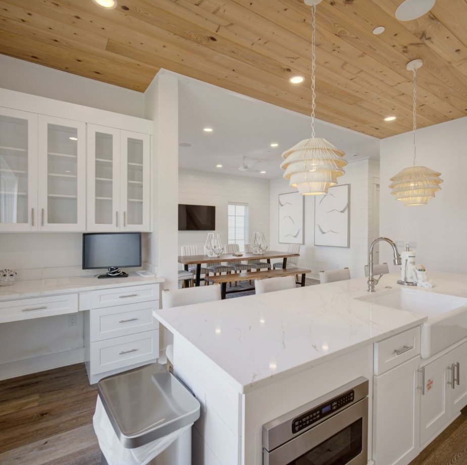 Modern kitchen with white cabinetry, marble countertops, built-in microwave, and a double sink, adjacent to a dining area with a wooden table and four chairs, decorated with two hanging pendant lights and wall art featuring abstract bird images.