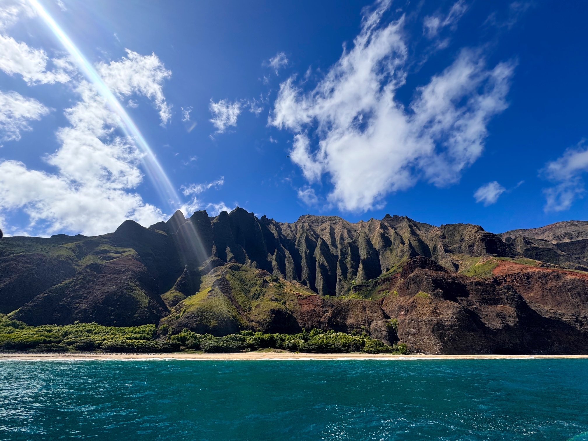 A landscape of a turquoise ocean with a sandy shoreline, lush green trees, mountainous terrain with dark rugged peaks, and a blue sky with some white clouds and a bright sun.