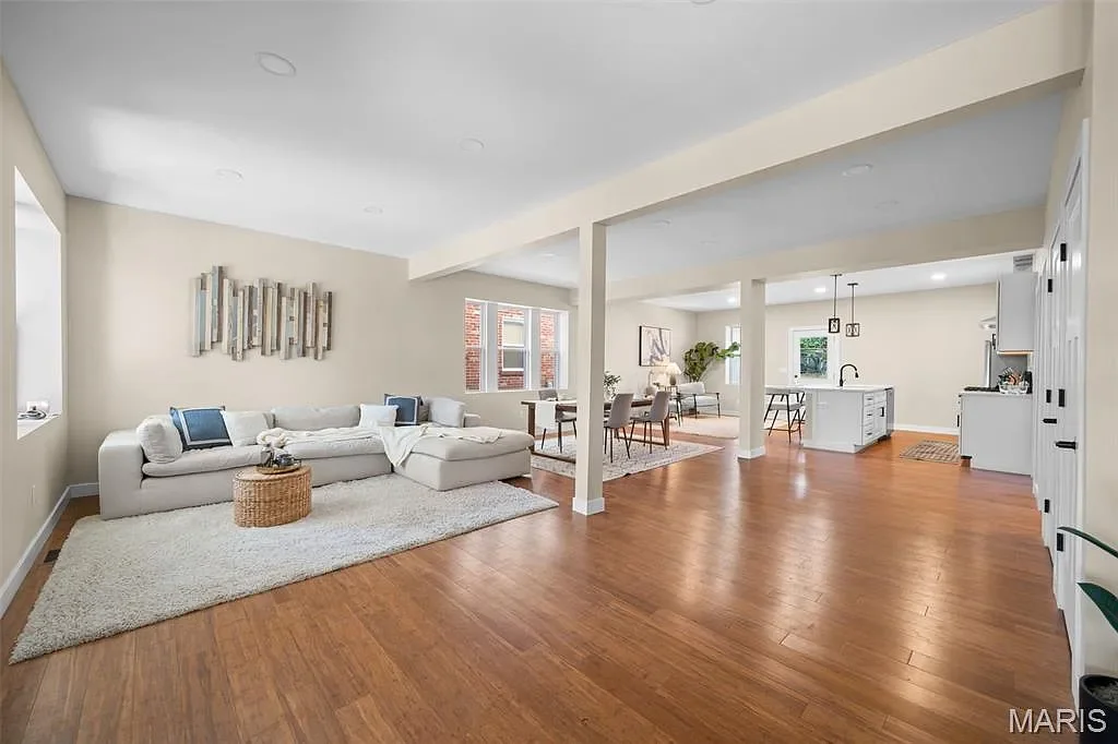 Open-concept living room, dining area, and kitchen with hardwood floors, white walls, and modern furniture. The living room has a white sofa with blue accent pillows and a textured shag rug. The dining area features a table with chairs, and the kitchen has white cabinetry, an island, and pendant lighting.
