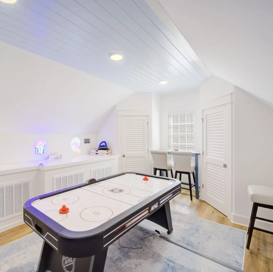 A room with white walls and ceiling, featuring a hockey table and neon jellyfish signs on the wall, along with a small seating area near a window.