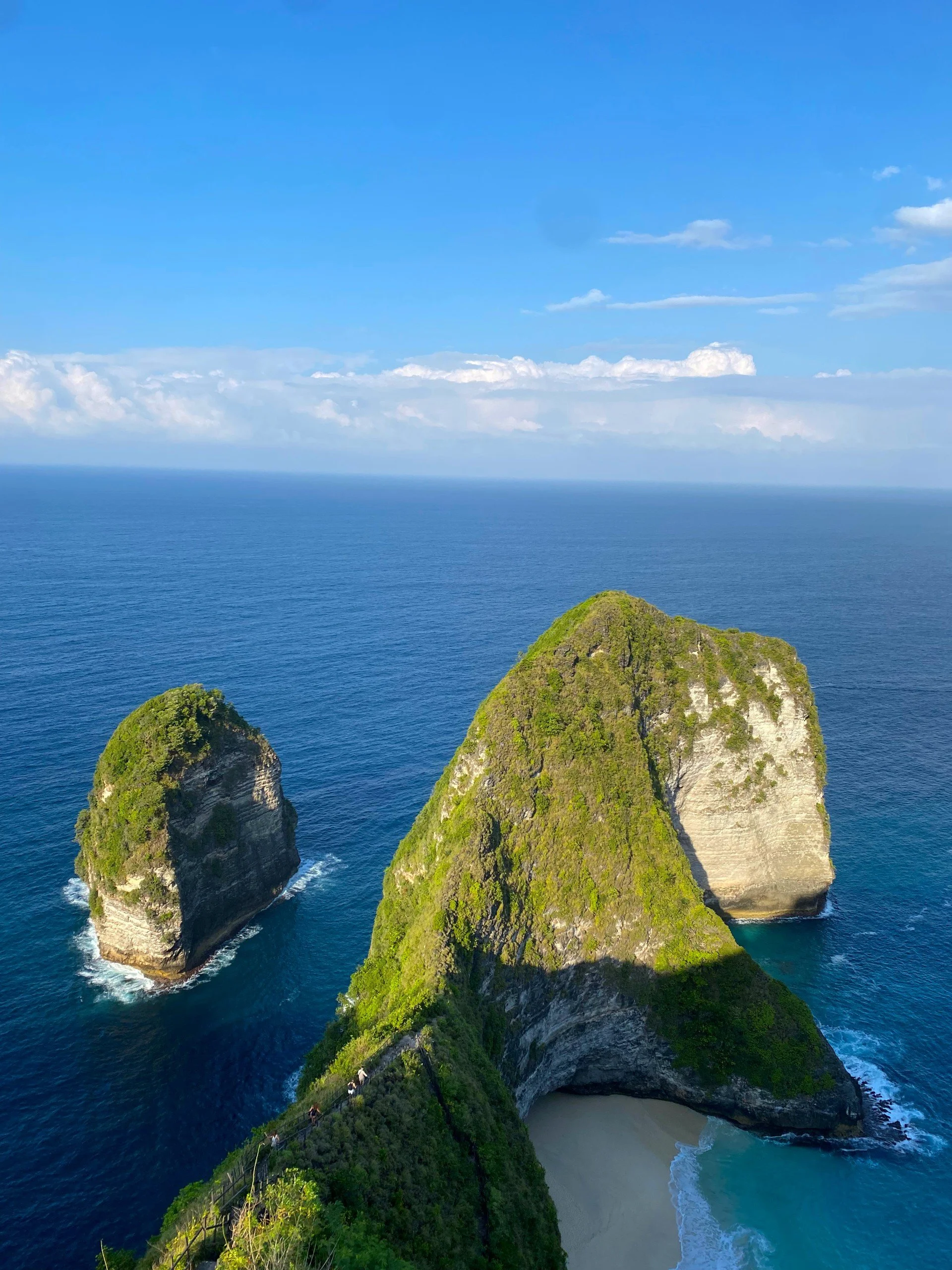 Three large, lush green cliffs rising from the ocean with a small sandy beach at their base, under a blue sky with scattered clouds.
