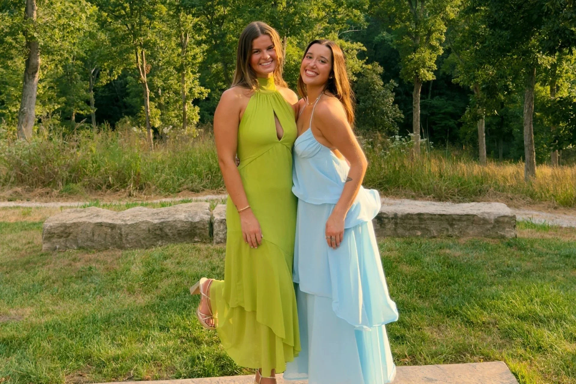 Two women standing together outdoors in a park, smiling at the camera, with trees and grass in the background.
