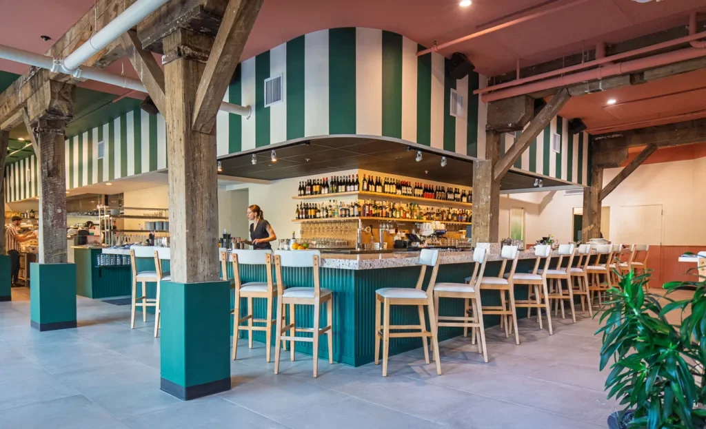 A bar inside a restaurant with a curved counter, bar stools, and shelves with bottles behind the counter. The decor features green and white striped walls, exposed wooden beams, and a pink ceiling.