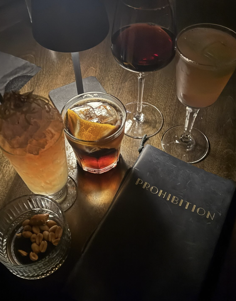 A dark wooden table with various drinks, a glass of red wine, a beige-colored cocktail, and a glass of dark soda with ice. There is also a glass of light-colored cocktail, a small glass of mixed nuts, a black book titled 'Prohibition,' and a black lamp.