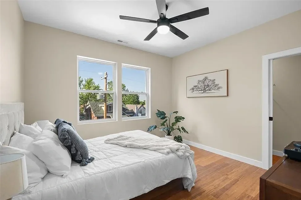 A bedroom with a white bed, beige walls, hardwood floor, a ceiling fan, two windows, a door, a framed tree artwork, and a potted plant.