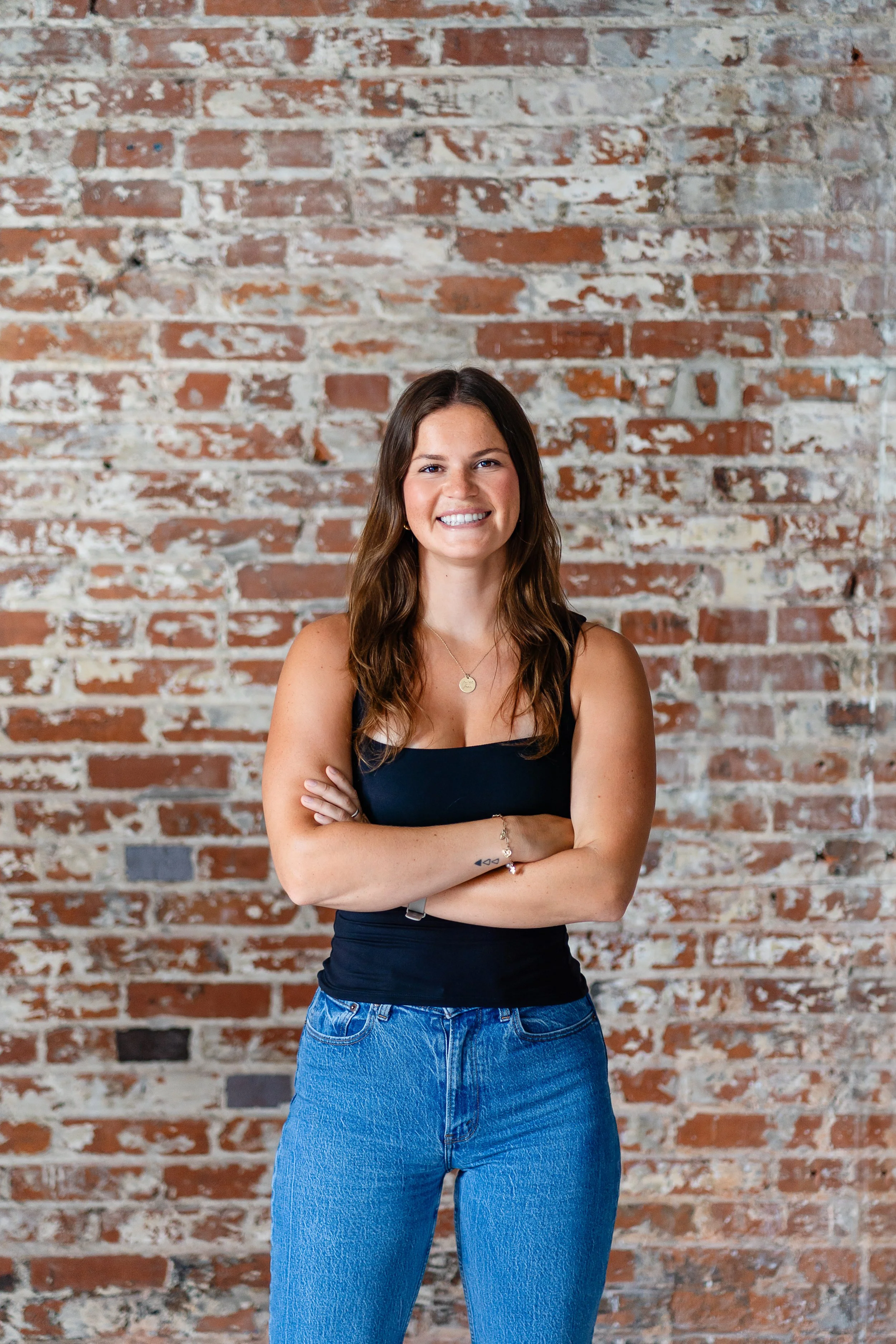 A young woman with long brown hair, wearing a black sleeveless top and blue jeans, standing with arms crossed in front of a brick wall.