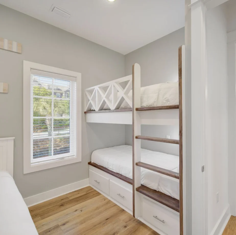 A small bedroom with a window, a white bunk bed with a built-in ladder, and light wood flooring.