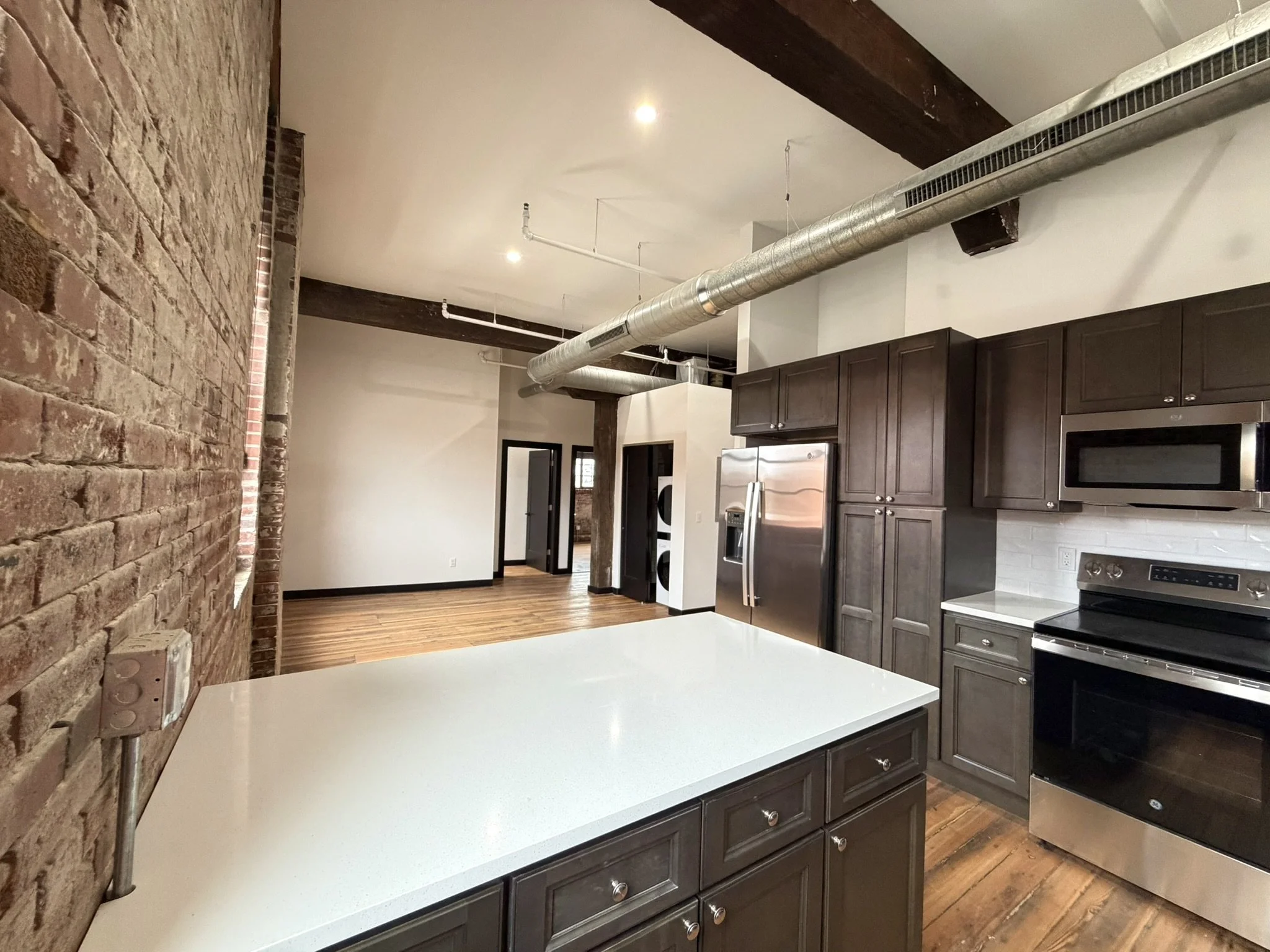 A kitchen with brick and white walls, dark wood cabinets, and stainless steel appliances. Exposed ductwork runs along the ceiling with wooden beams.