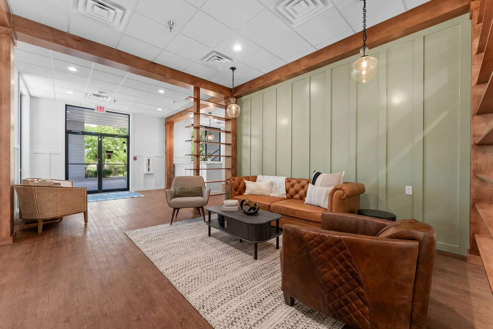 A modern lobby with wooden accents, leather and fabric chairs, a green paneled wall, pendant lights, and a front door with glass panels and an exit sign.