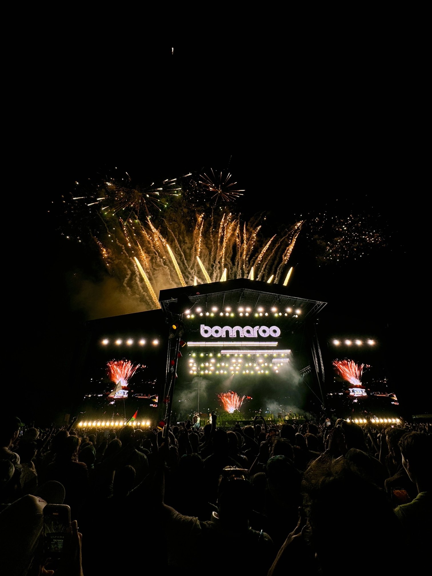 Crowd watching a fireworks display at night with a stage that has a banner reading 'bomaaro' in the foreground.