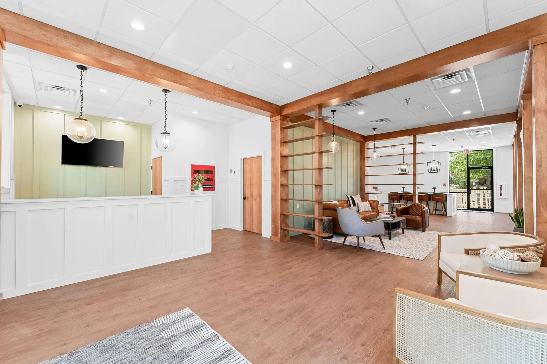 Interior of a modern lounge or waiting area with wood flooring, white walls, and a ceiling with recessed lighting and hanging globe lights. The space includes a seating area with a gray armchair, a brown leather sofa, and a wooden coffee table, separated by wooden structural dividers. Large window and glass door at the back provide natural light and outdoor view.