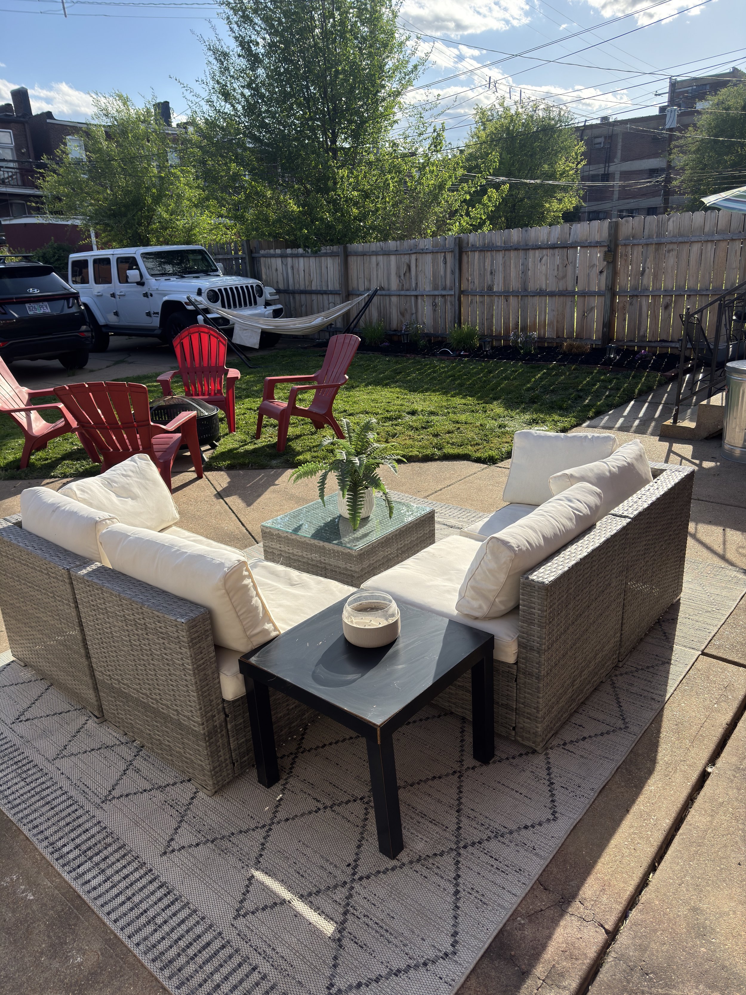 A backyard patio with outdoor furniture, including a wicker sofa with white cushions, a black side table, and a potted plant on the table. There are red and pink plastic chairs and a small black firepit on a patterned mat. In the background, a wooden fence separates the yard from neighboring buildings, with a white Jeep parked in front. An outdoor hammock hangs between two trees, and the sky above is partly cloudy with sunlight casting shadows.