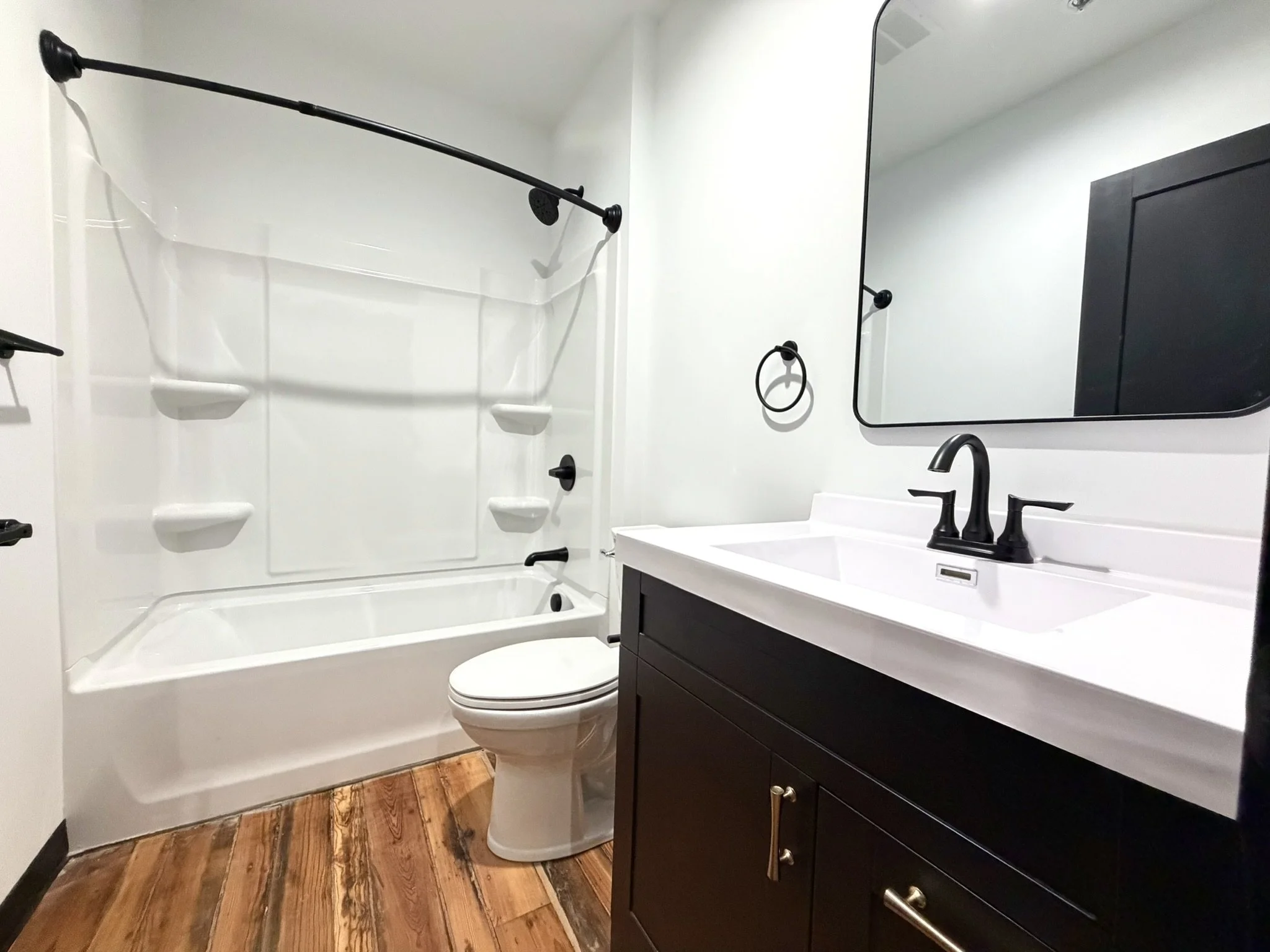 A modern bathroom with a bathtub and shower combination with black fixtures and white walls, a dark wood vanity with a white countertop and black faucet, a large mirror, a black towel ring, a toilet, and hardwood flooring.
