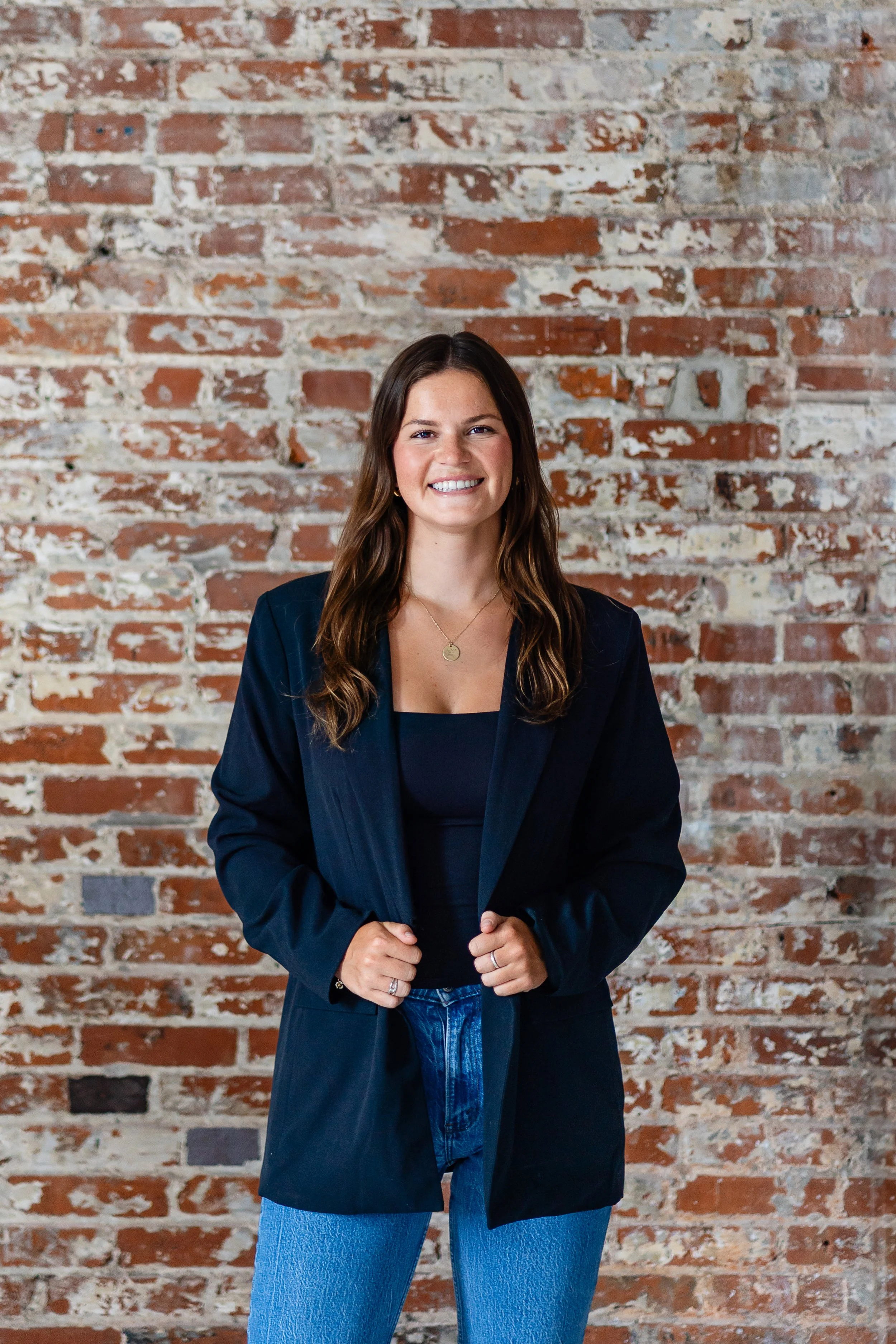 A smiling woman with long brown hair wearing a navy blazer, black top, and blue jeans standing in front of a rustic brick wall.