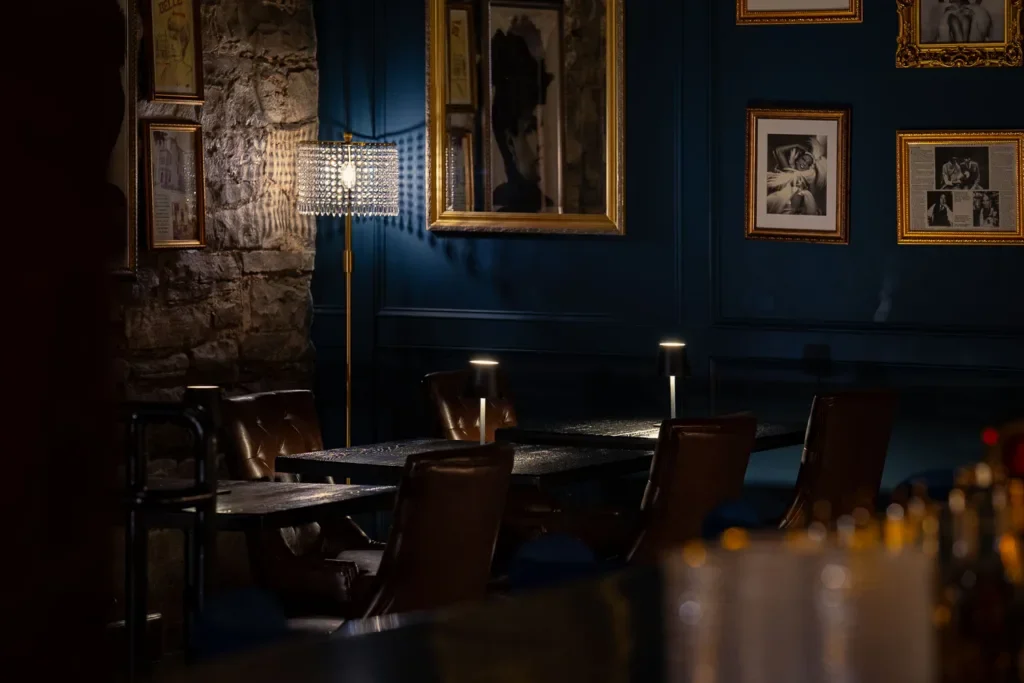 Interior of a dimly lit dining area with dark blue walls, framed black-and-white photographs, a stone wall, and a tall floor lamp with a beaded lampshade, along with several chairs around tables.