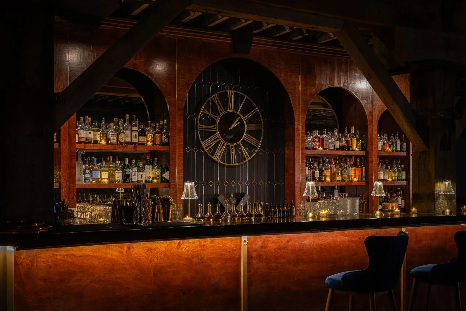 A dimly lit bar with wooden shelves filled with various liquor bottles, a large clock on the wall, and small lamps on the counter, with two dark chairs in front.