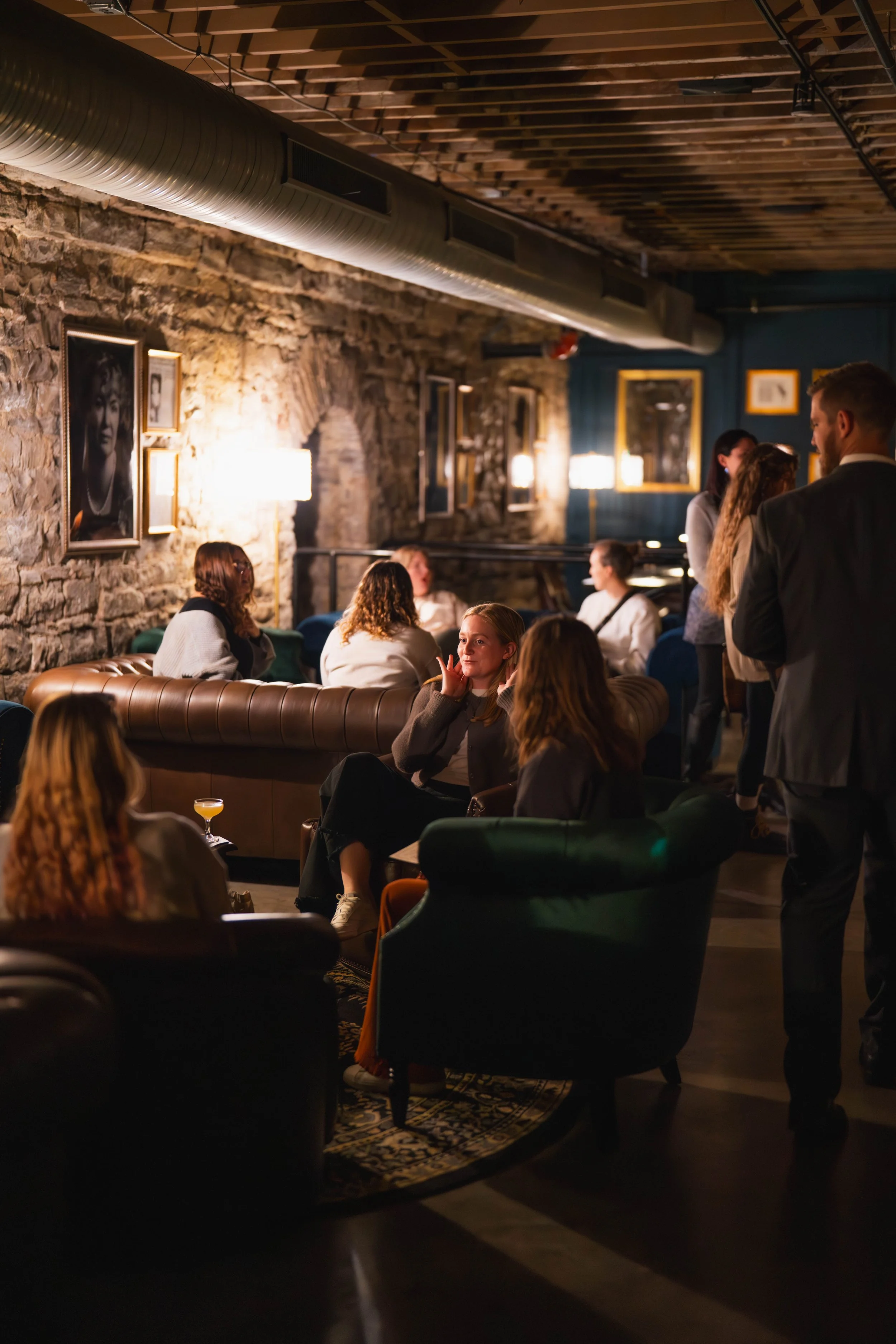 People gathered in a cozy, dimly-lit lounge or restaurant with exposed brick walls, framed photographs, and vintage-style lighting, engaged in conversations.