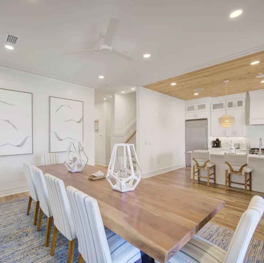 Open-concept dining and kitchen area with wooden flooring, white walls, and modern decor. The dining table has eight striped chairs, and the kitchen features white cabinets, stainless steel appliances, and two bar stools at the counter. Recessed lighting and a ceiling fan are visible.