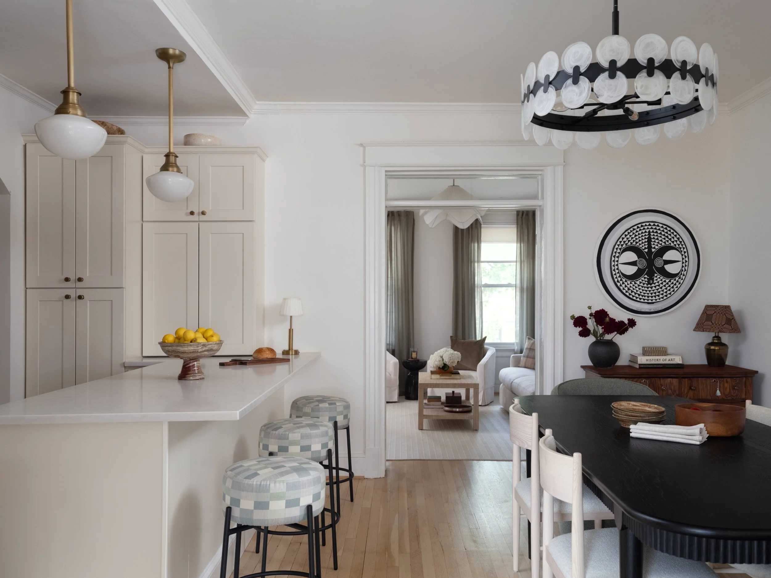 A modern kitchen and living room with white cabinetry, pendant and chandelier lighting, a window with curtains, a round black dining table with white chairs, a black and white wall art, and various decorative items including a bowl of lemons and a fl