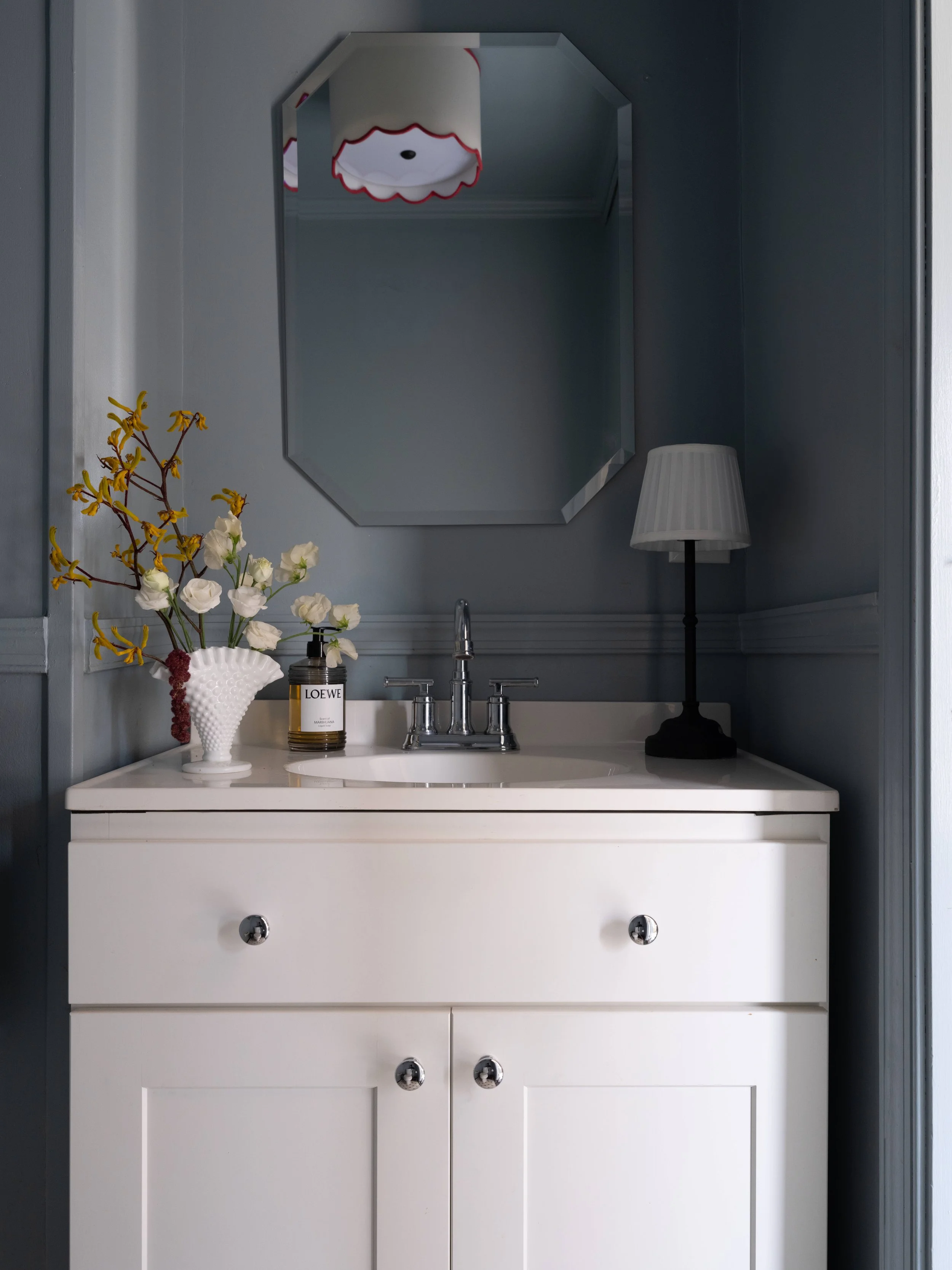 A white bathroom vanity with a silver faucet, a vase with white and yellow flowers, a black and white candle labeled 'LOEWE', a black table lamp with a white lampshade, a large rectangular mirror, and a ceiling light fixture, set against dark walls.