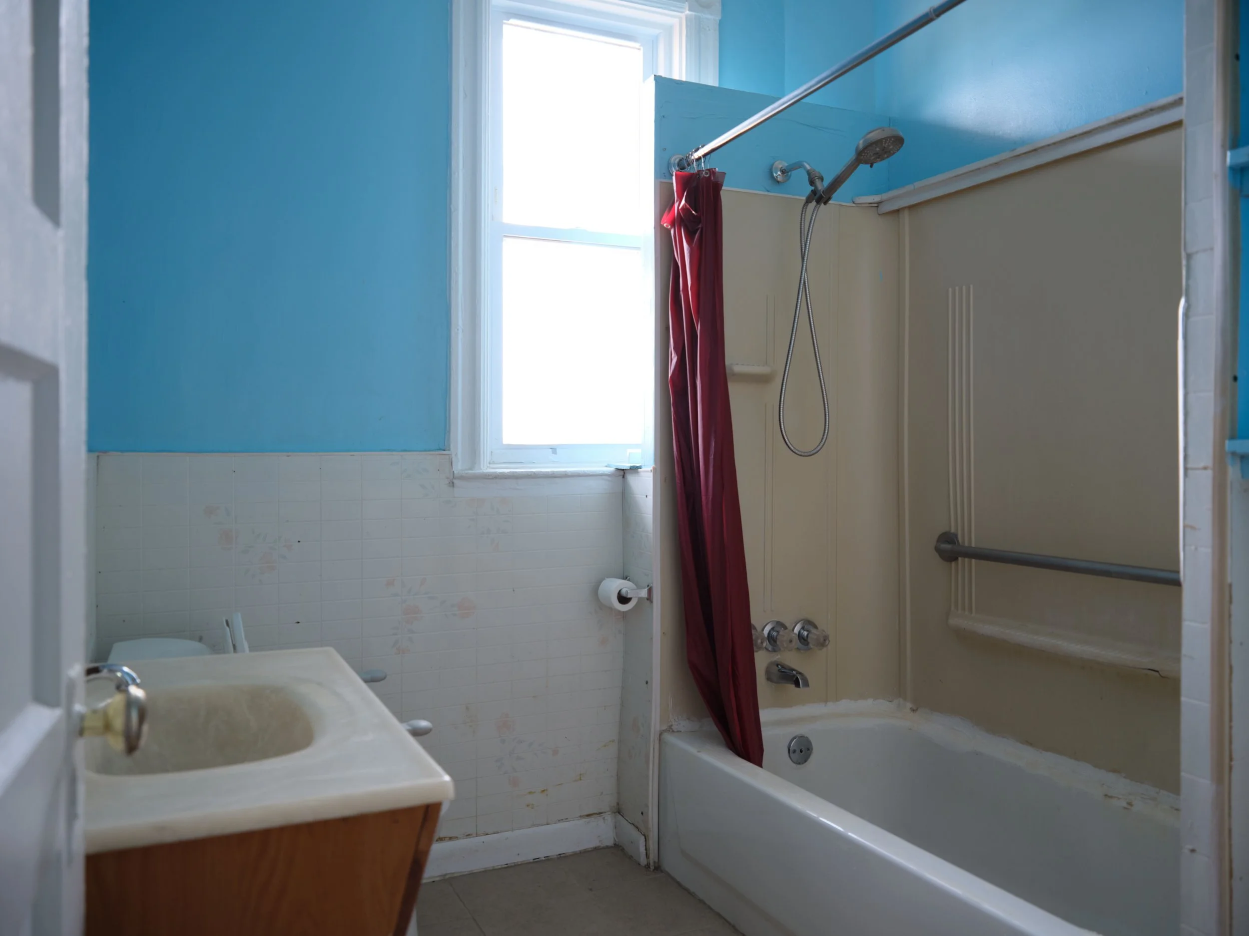 A bathroom with blue walls, a bathtub with a red shower curtain, a window letting in natural light, a white sink, and a toilet. The bathtub has a shower head and fixtures.