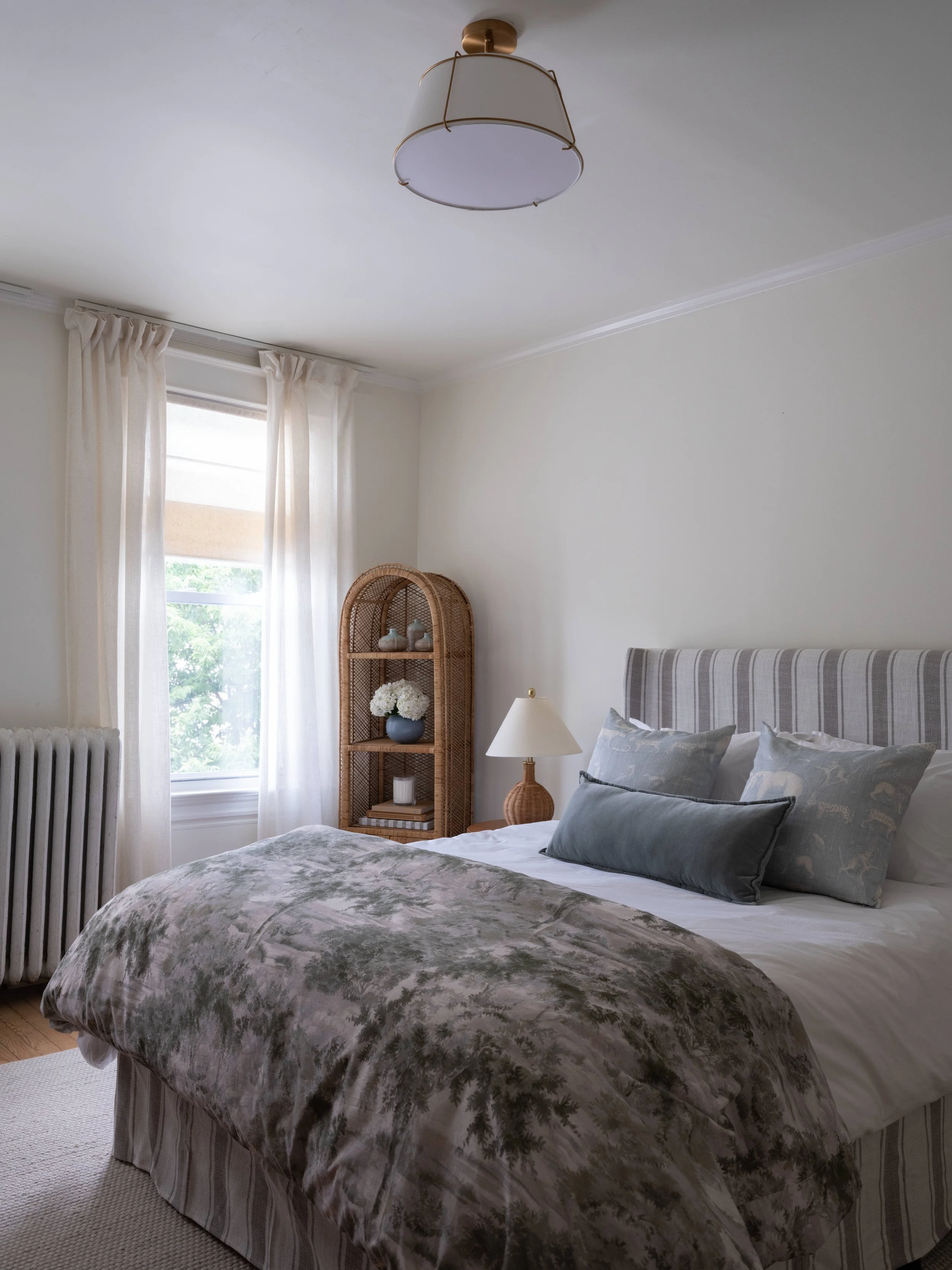 A cozy bedroom with a large bed covered in a floral comforter and decorative pillows, near a window with white curtains, and a rattan shelving unit with decorative items and a vase of flowers.