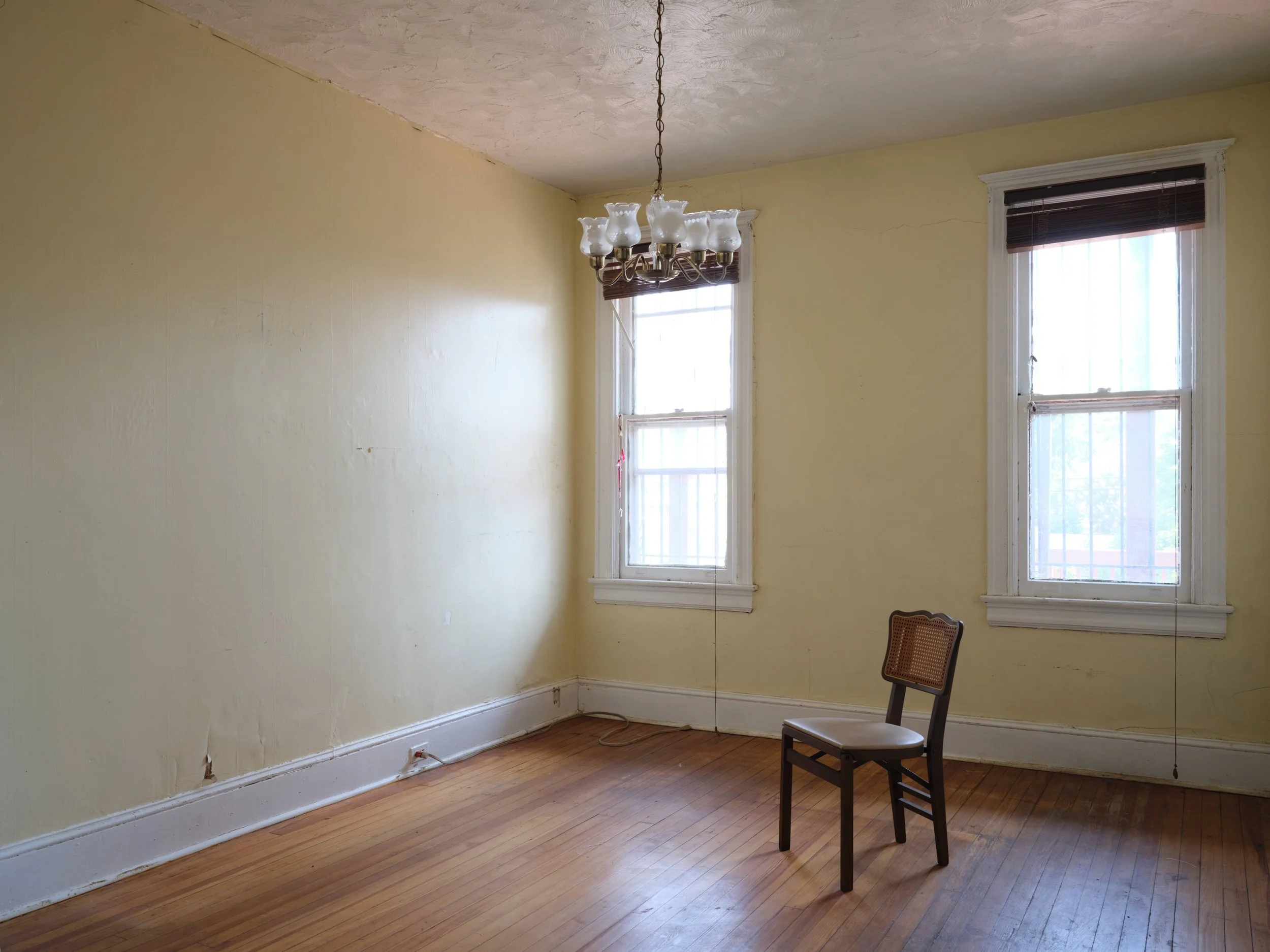 Empty room with yellow painted walls, two large windows with dark blinds, a wooden floor, a ceiling light fixture, and a single chair with a woven backrest.