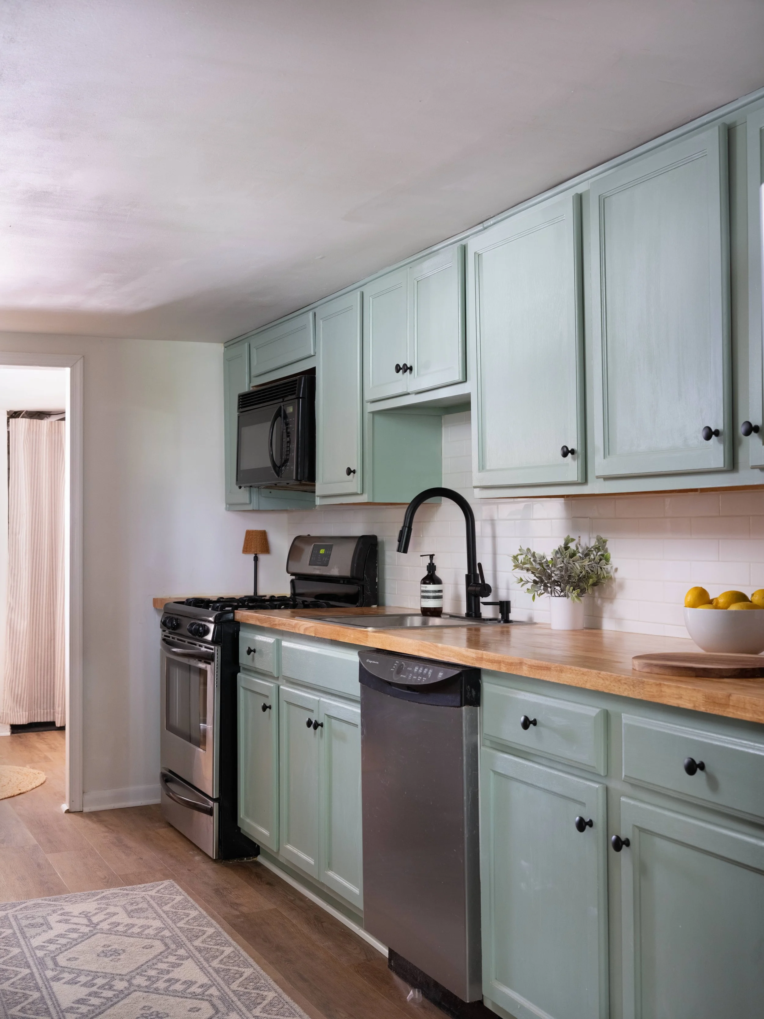 A kitchen with pastel green cabinets, a black microwave, a stove, and a stainless steel dishwasher. The countertop has a potted plant and a bowl of lemons. The floor is wood, and there's a patterned rug near the doorway.