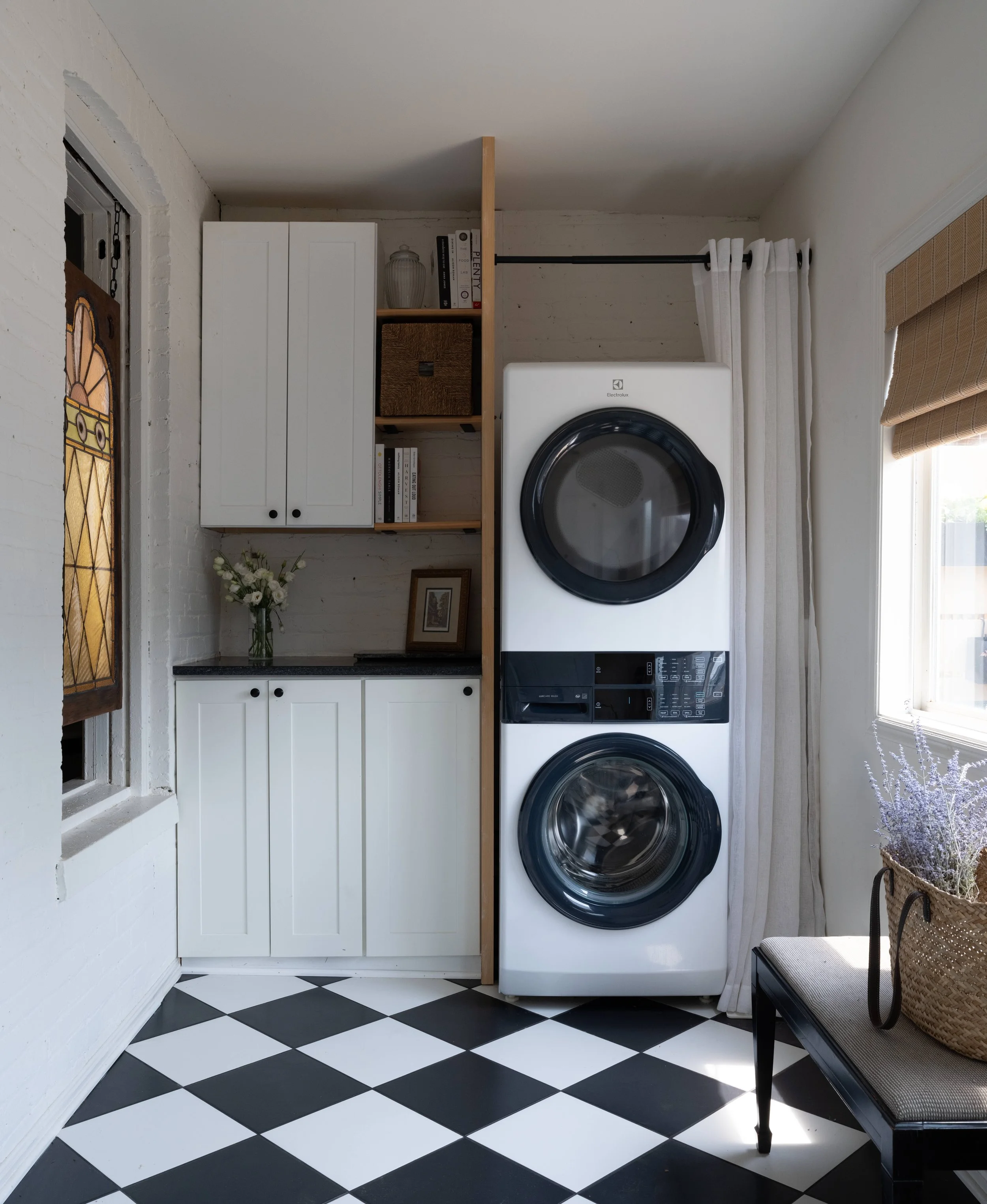 A laundry room with black and white diamond checkered floor, white brick walls, white cabinets, a washing machine, a dryer, a woven storage basket, a vase with white flowers, a framed picture, a black bench with a straw bag, and a window with beige Roman shades.