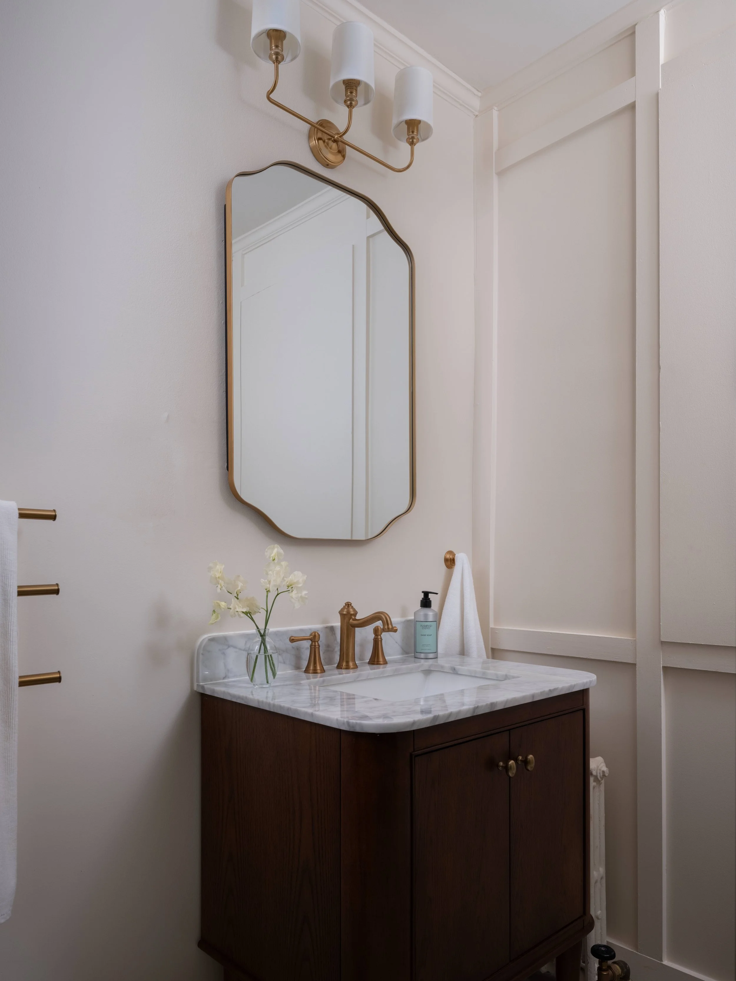 Bathroom vanity with dark wood cabinet, white marble countertop, gold faucet, a vase with white flowers, green soap dispenser, mirror, and wall light fixture with three white shades.