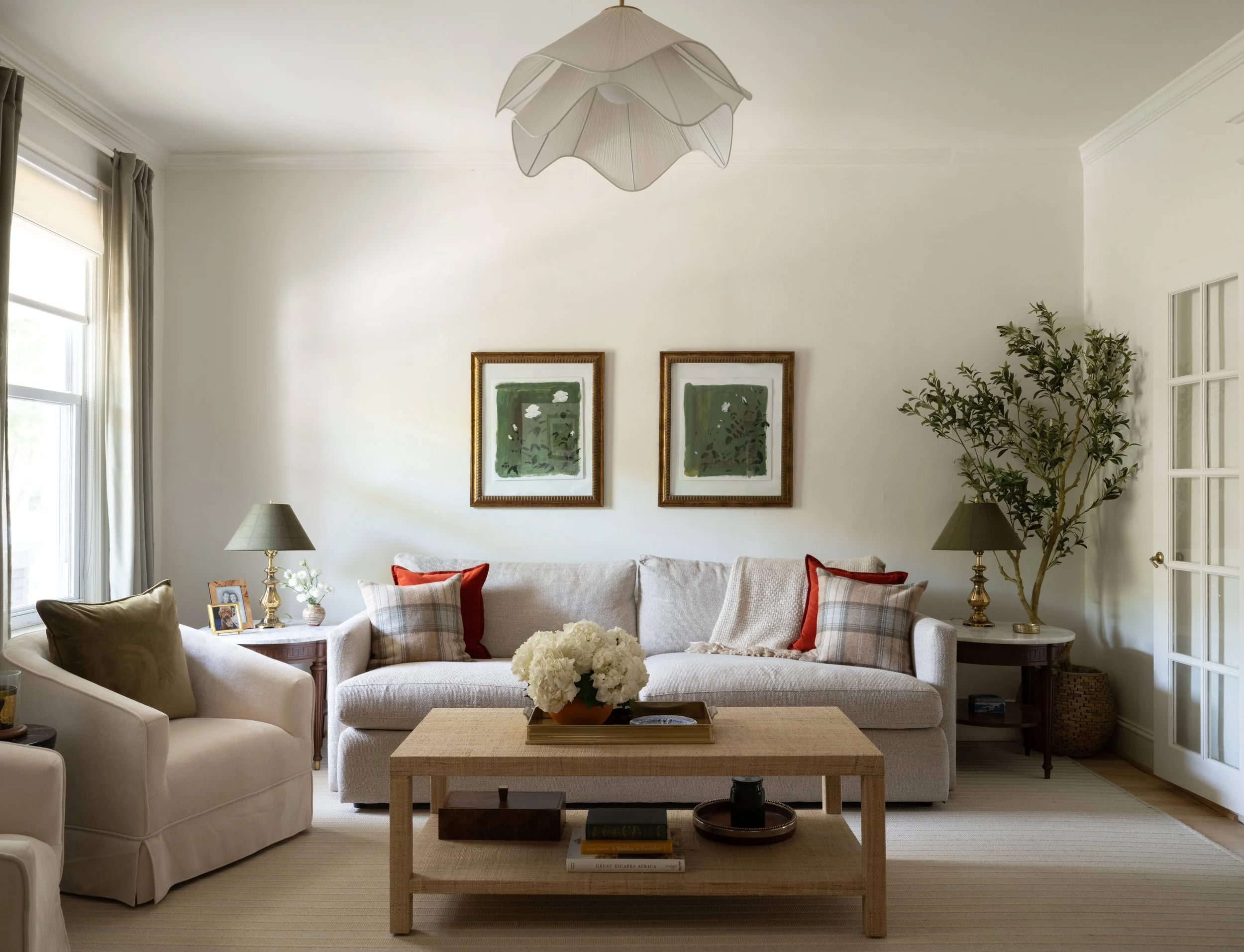 Living room with white sofas, framed artwork, and a wooden coffee table with a flower arrangement, large windows with beige curtains, and a ceiling light fixture.