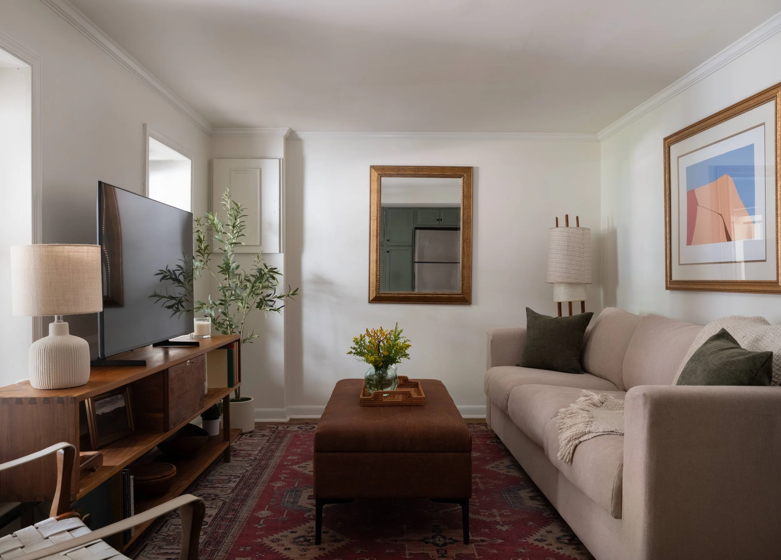 Living room with a beige sofa, a wooden ottoman, a TV on a stand, a plant, wall art, a floor lamp, and a mirror
