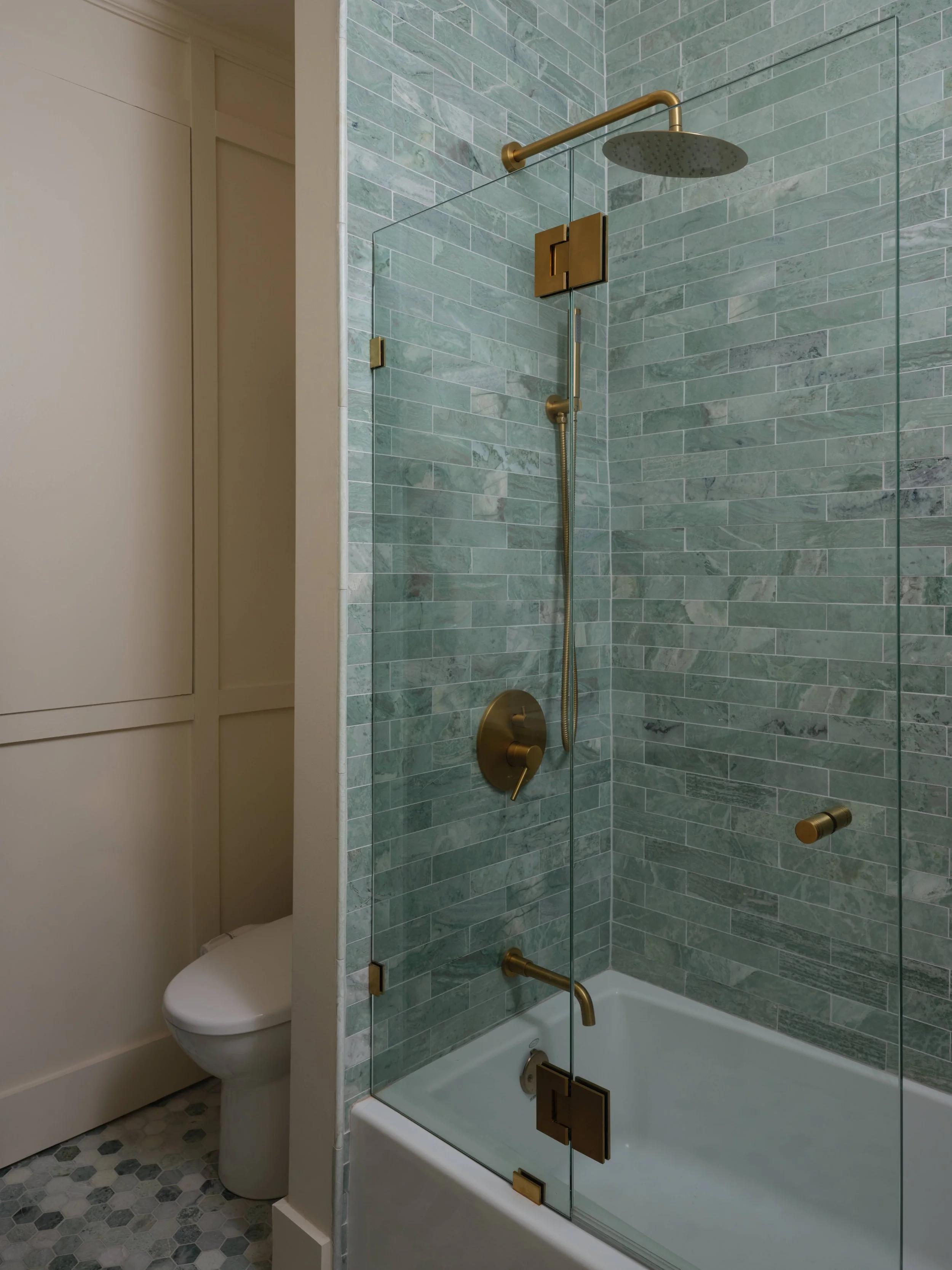 Bathroom with green tiled wall, brass shower fixtures, glass shower enclosure, and a hidden toilet behind a partition.