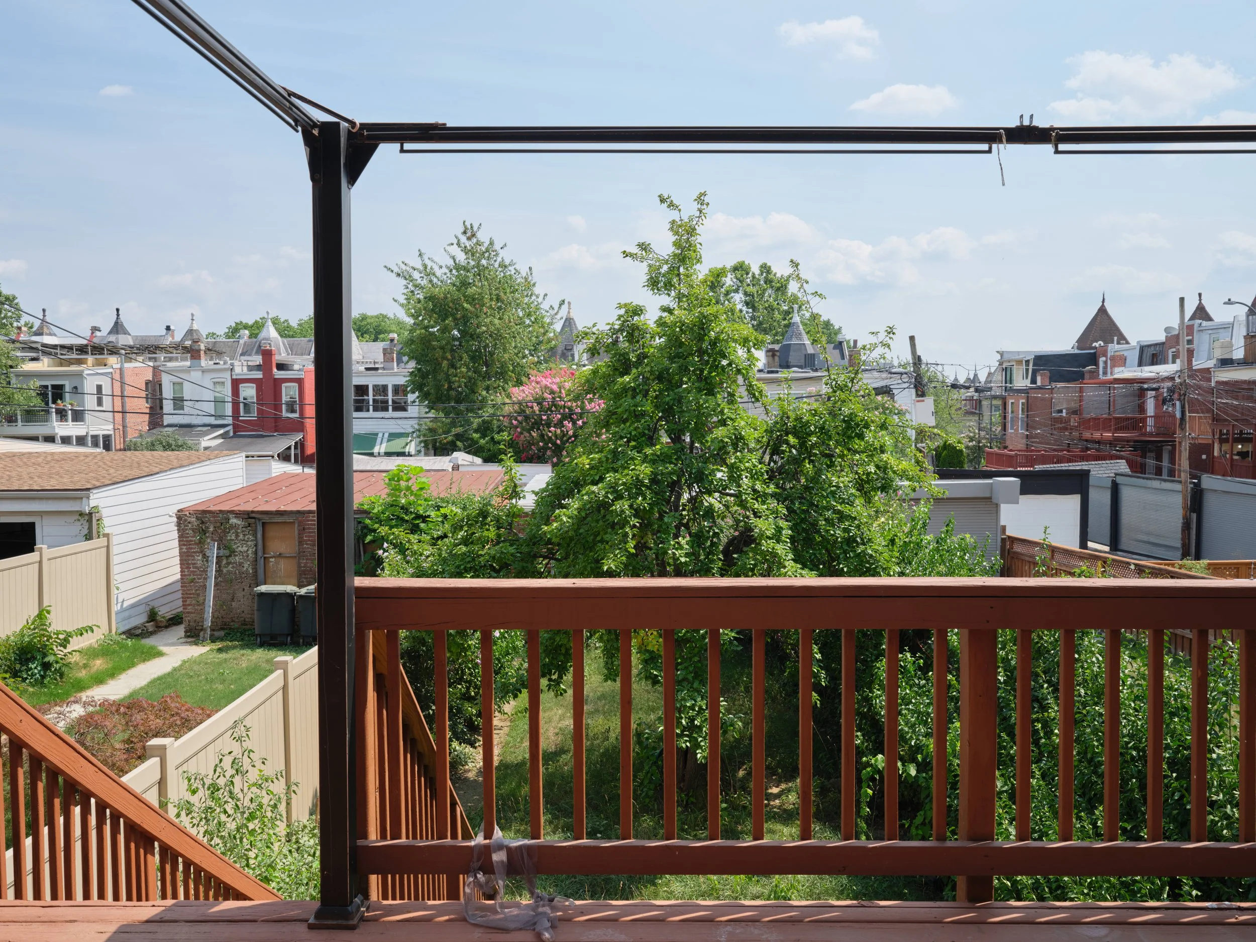 View from a wooden balcony overlooking suburban houses with rooftops, trees, and a blue sky with scattered clouds.