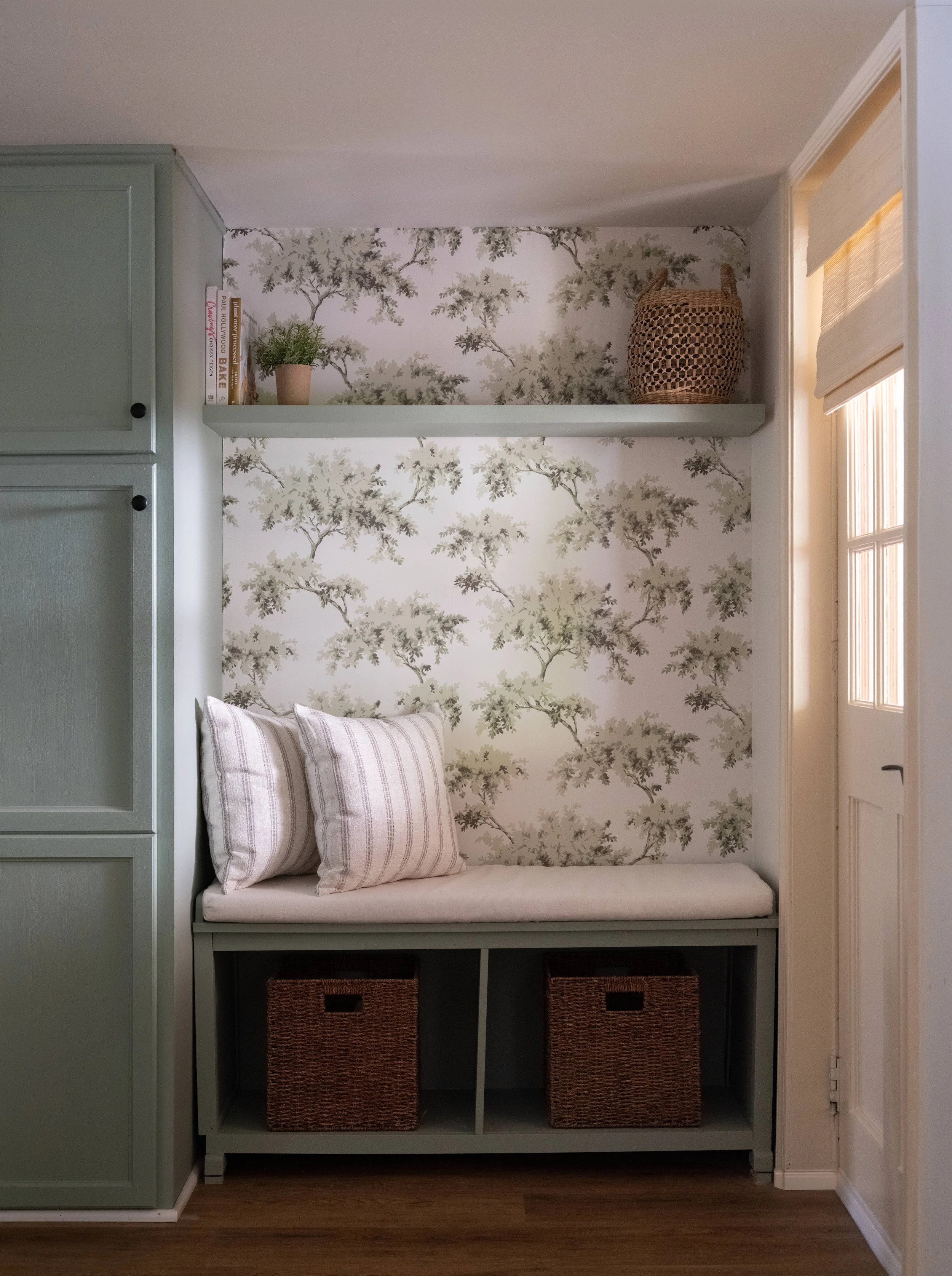 A built-in seating nook with a cushioned bench and two striped pillows, set against a wall with tree-patterned wallpaper. Above the nook, a shelf holds books, a potted plant, and a woven basket. To the left, part of a gray cabinet is visible, and to 