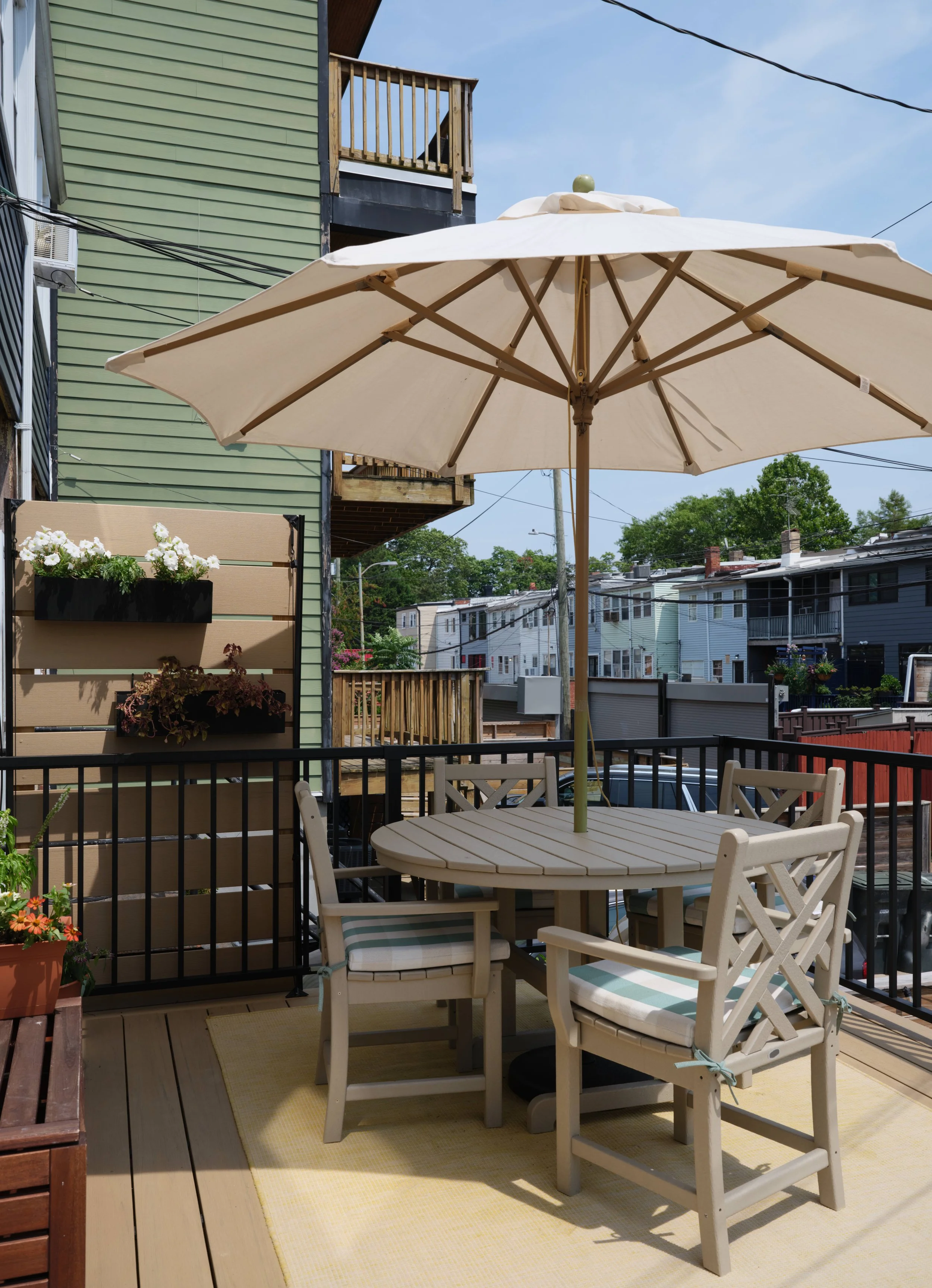 Outdoor balcony with a round white table, four white chairs with striped cushions, a large beige patio umbrella, potted flowers on a wall-mounted planter, and neighboring houses in the background under a blue sky.