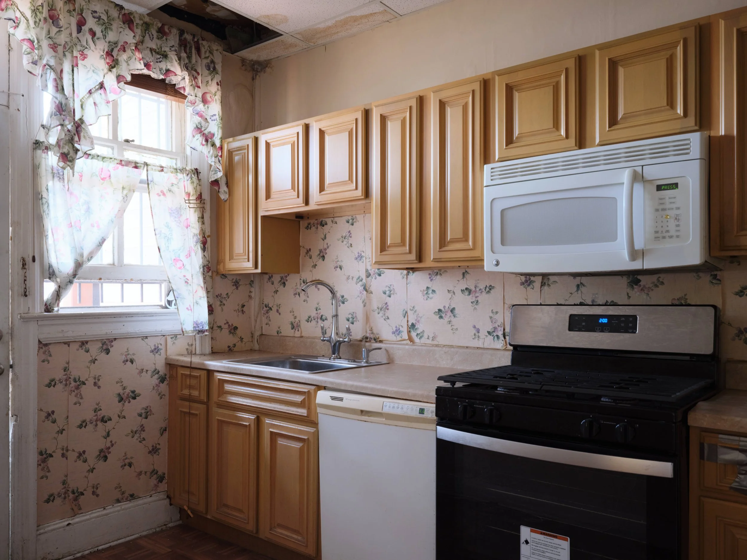 A kitchen with wooden cabinets, floral wallpaper, a window with floral curtains, a white microwave, and a black stove.