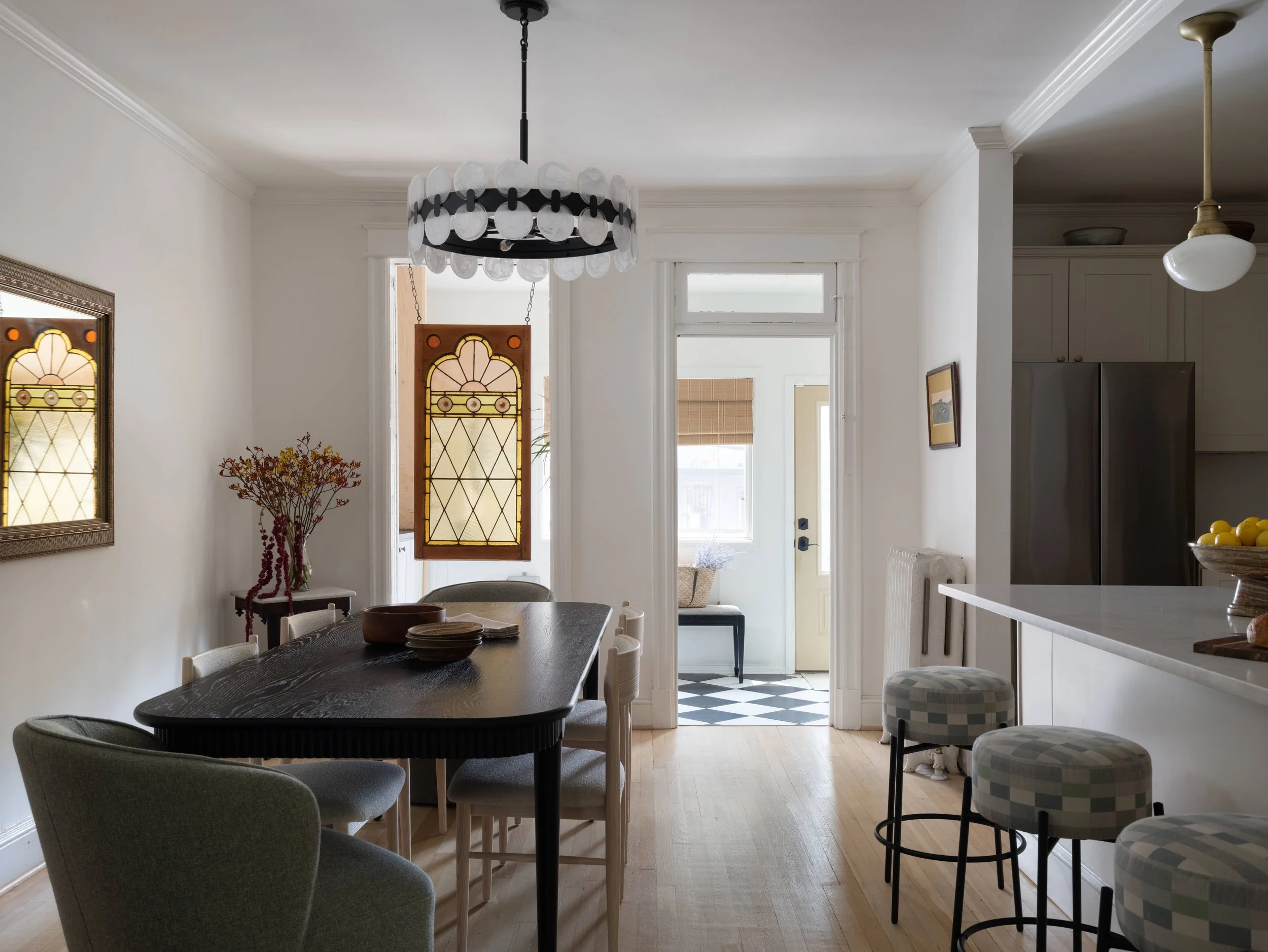 A dining area with a black table surrounded by light gray chairs, decorative stained glass doors, a ceiling chandelier made of clear glass discs, and a white ceiling and walls with crown molding. There's a vase with dried flowers on a small table, an