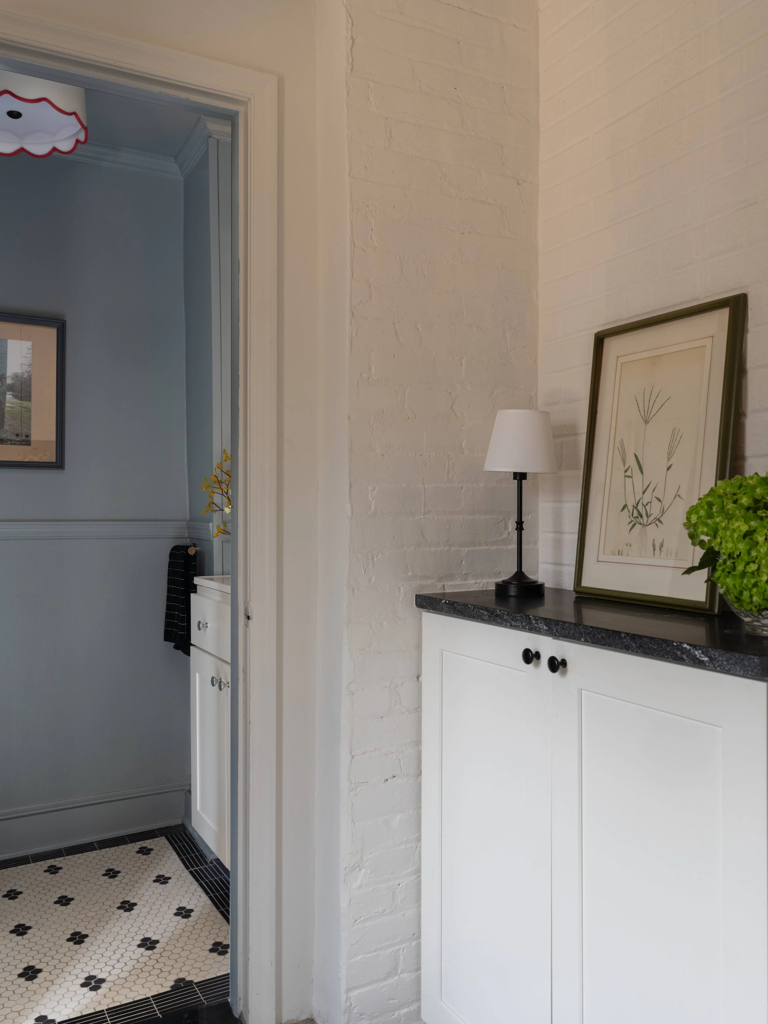 Interior of a room with white brick wall and white cabinet with black knobs, framed botanical art leaning against wall, white lamp on black countertop, partial view of a bathroom with blue walls and black and white tiled floor.