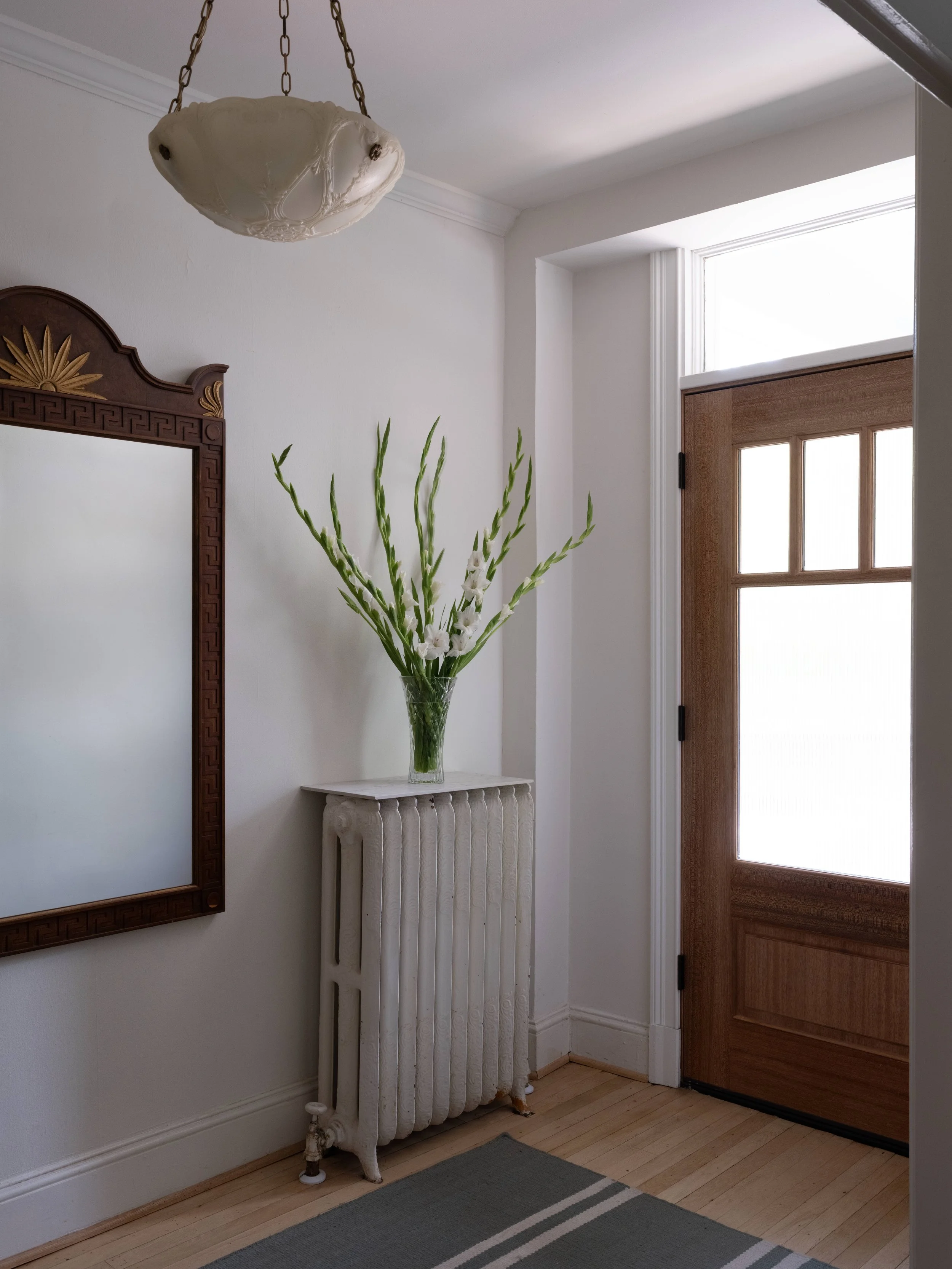 An entryway with a wooden door, a vintage radiator with a glass vase containing tall white gladiolus flowers on top, a wooden-framed mirror, and a gray rug on a light wooden floor.