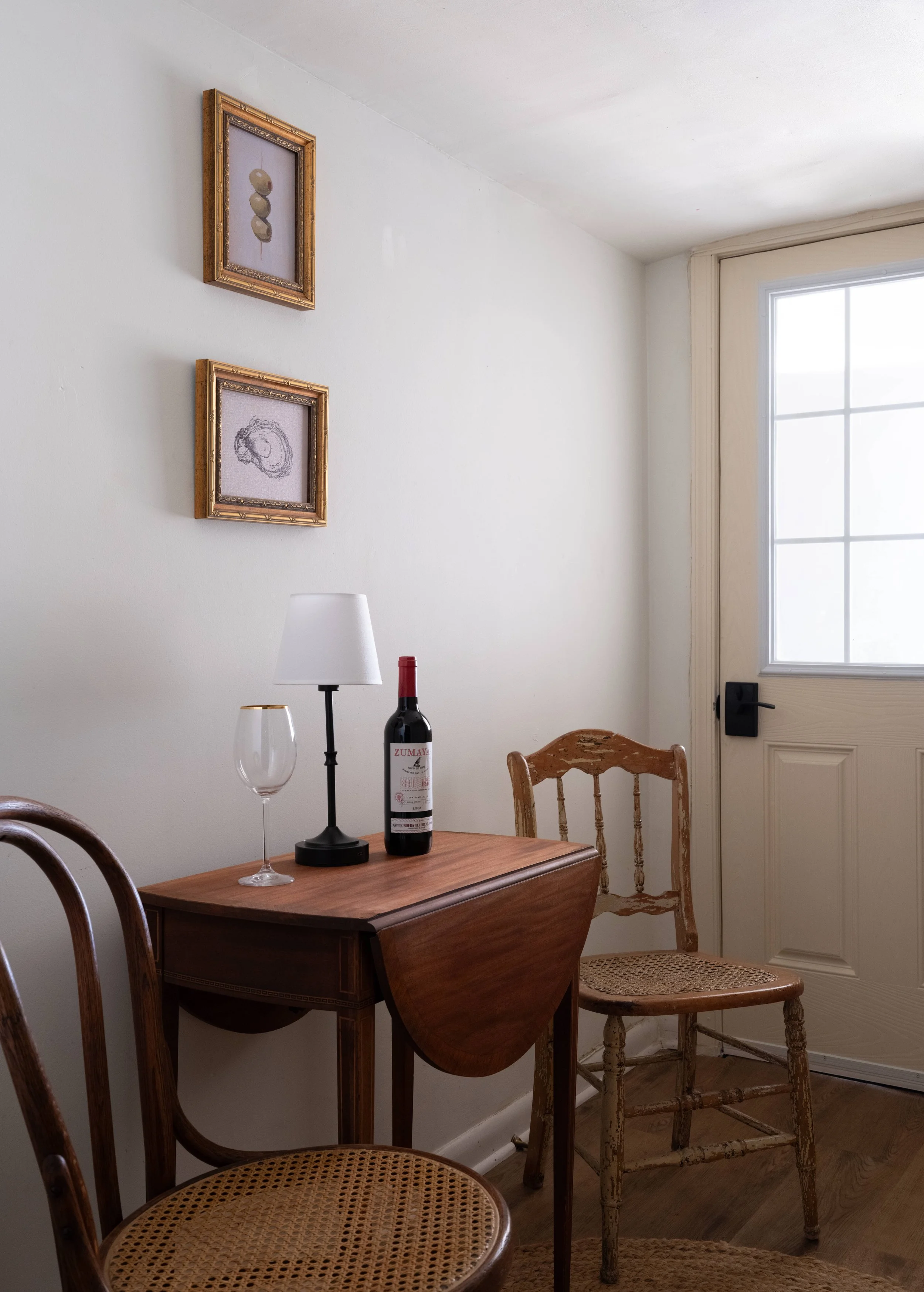 A corner of a room with a wooden dining table, several wooden chairs with cane seats, a white wall, a window with a beige door frame, framed art on the wall, and a table lamp with a wine bottle and filled wine glass.