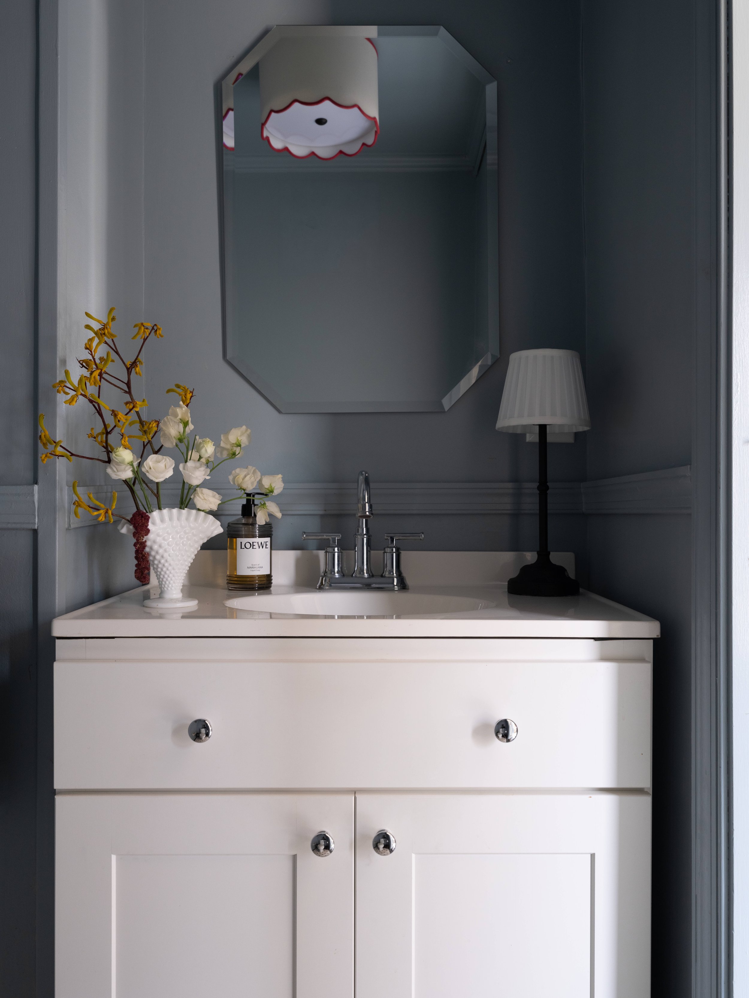 A white dresser with metallic knobs in a room with blue walls, decorated with a white vase of flowers, a bottle of Loewe perfume, a mirror, a lamp, and a sink faucet visible at the mirror's reflection.